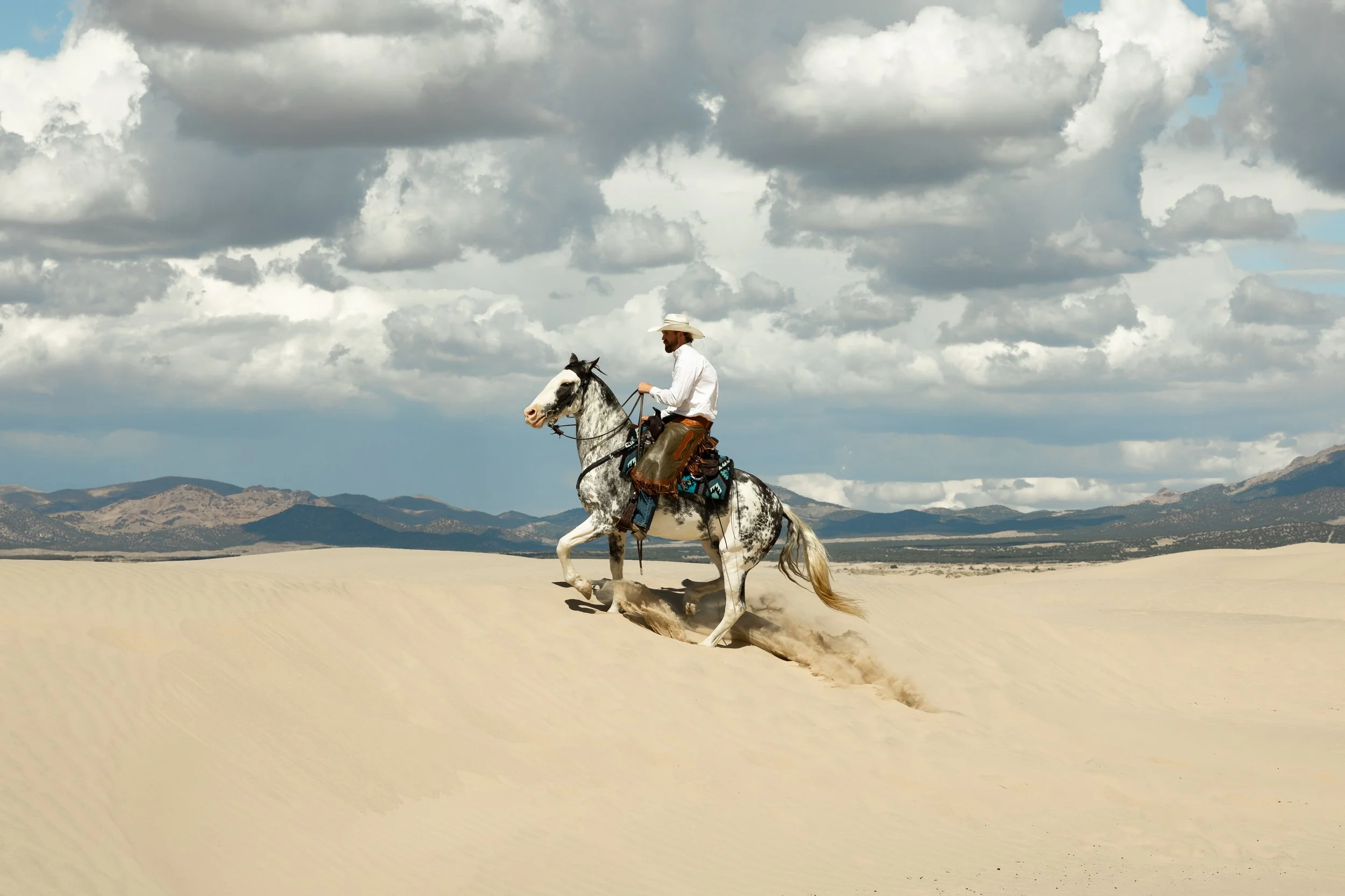 A man riding a white and gray horse across sandy desert dunes under cloudy sky.