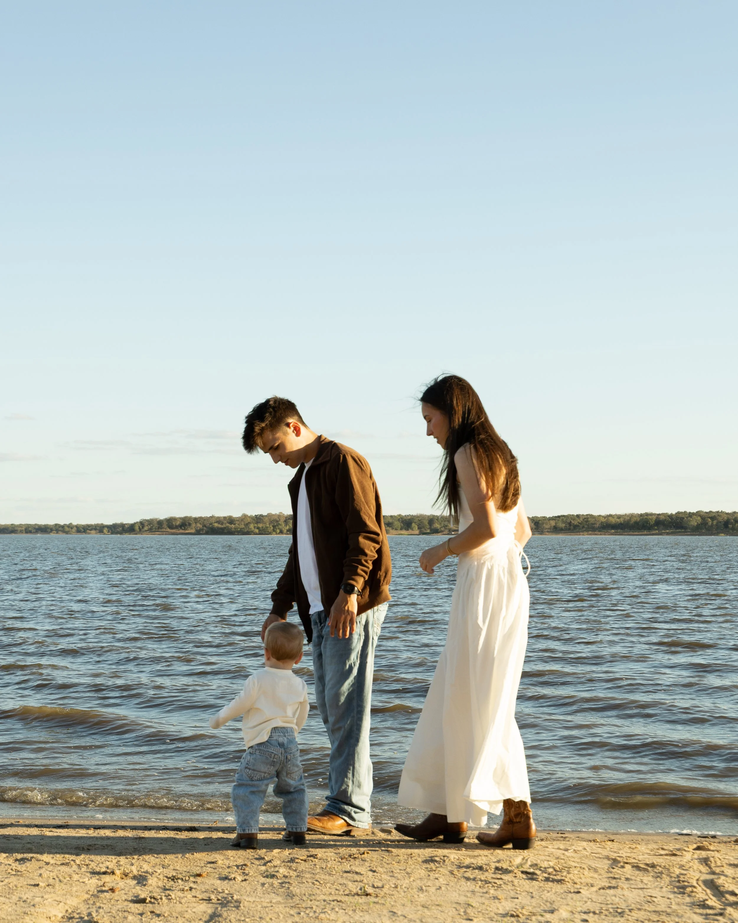 A family of three standing on a sandy beach by a lake, with the parents and a toddler looking at the water during sunset.