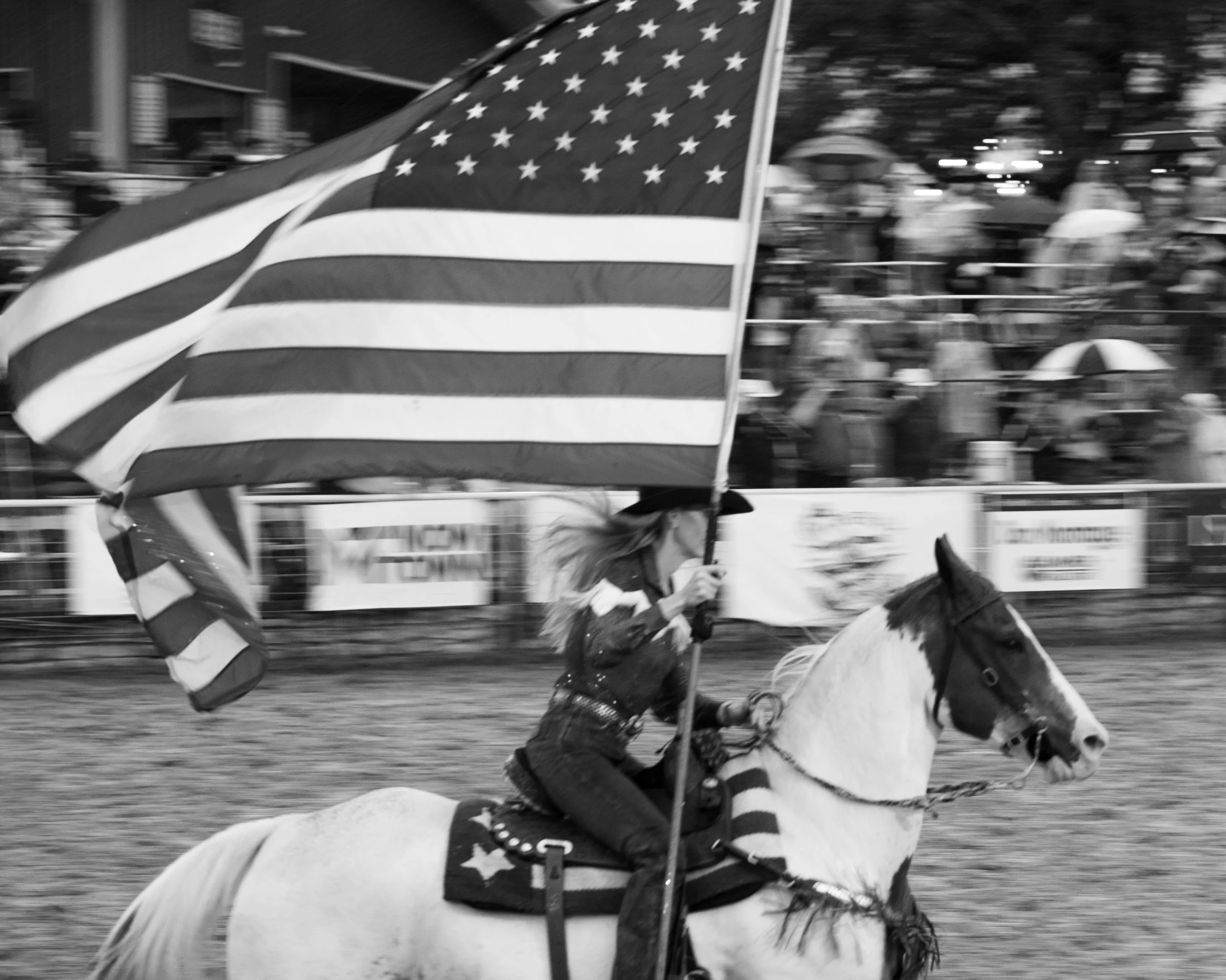 A woman riding a horse at a rodeo, holding an American flag with a crowd watching in the background.