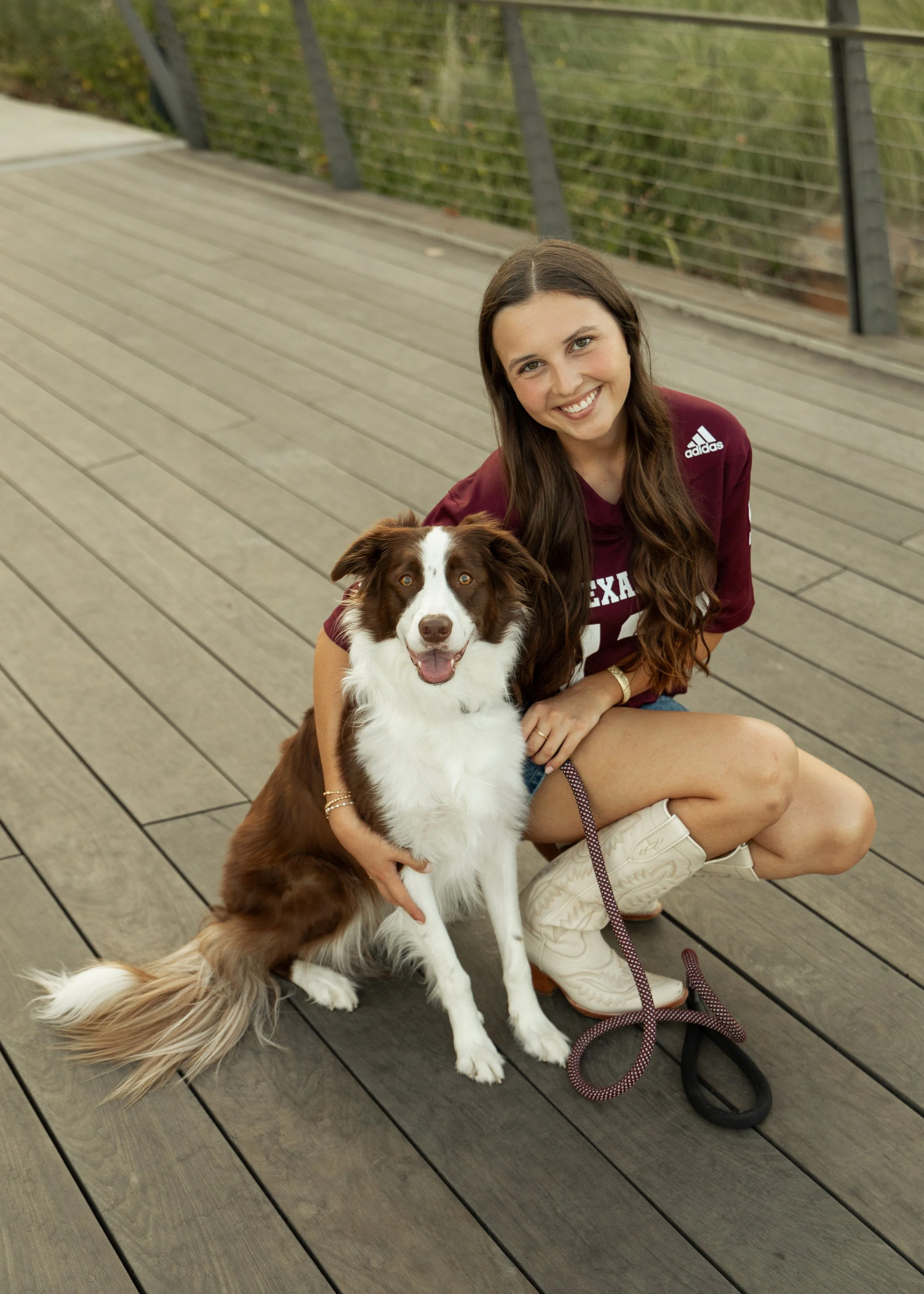 A young woman with long brown hair and a maroon Texas A&M shirt crouches next to a brown and white dog on a wooden bridge, both smiling at the camera.