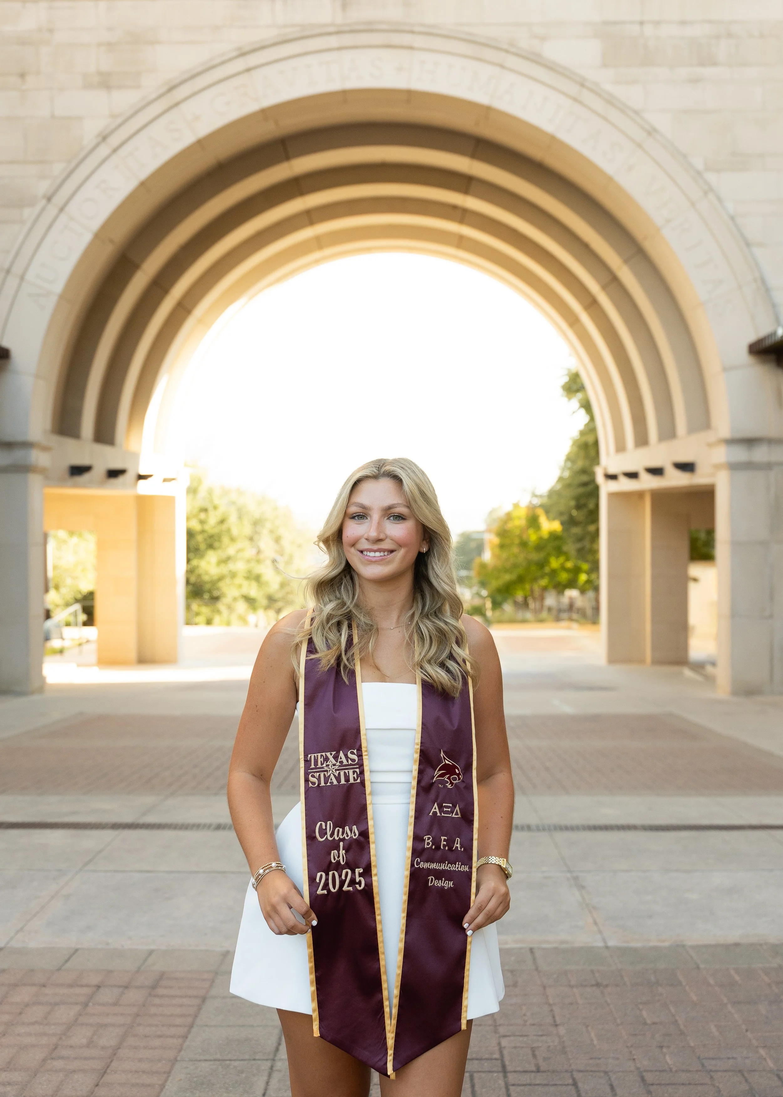 A young woman with blonde hair wearing a white dress and a maroon graduation stole, standing outdoors in front of an arched stone gateway, smiling at the camera.