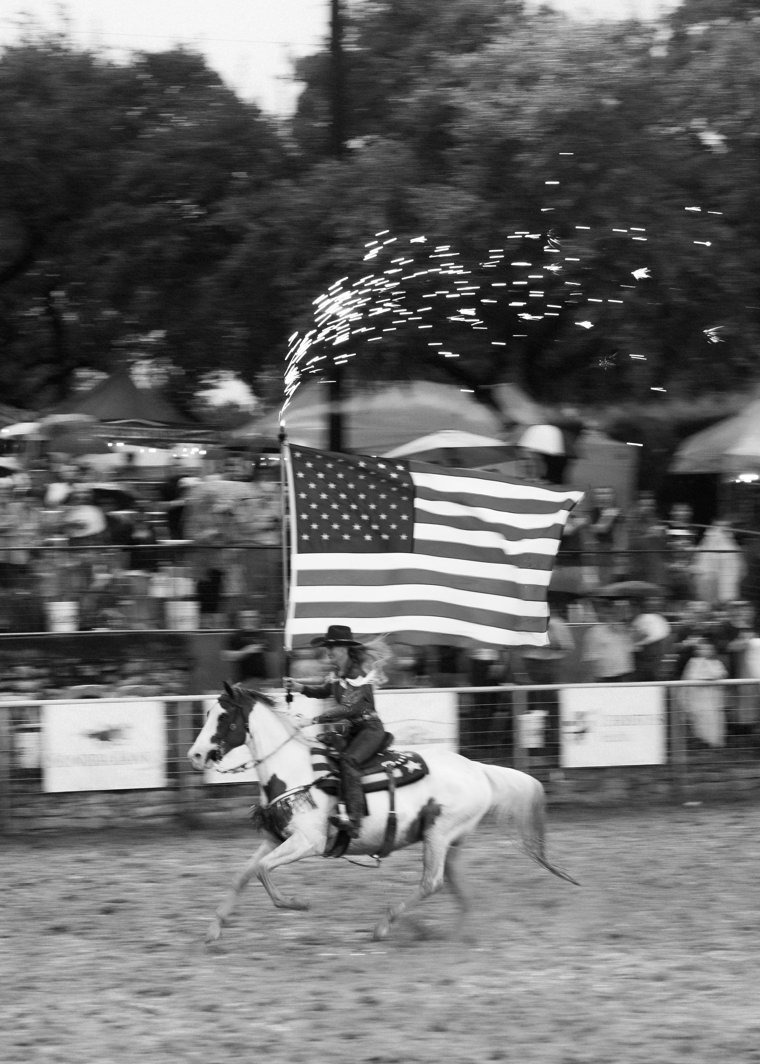 A young girl riding a horse at a rodeo or fair, holding a large American flag with fireworks in the background, black and white photo.