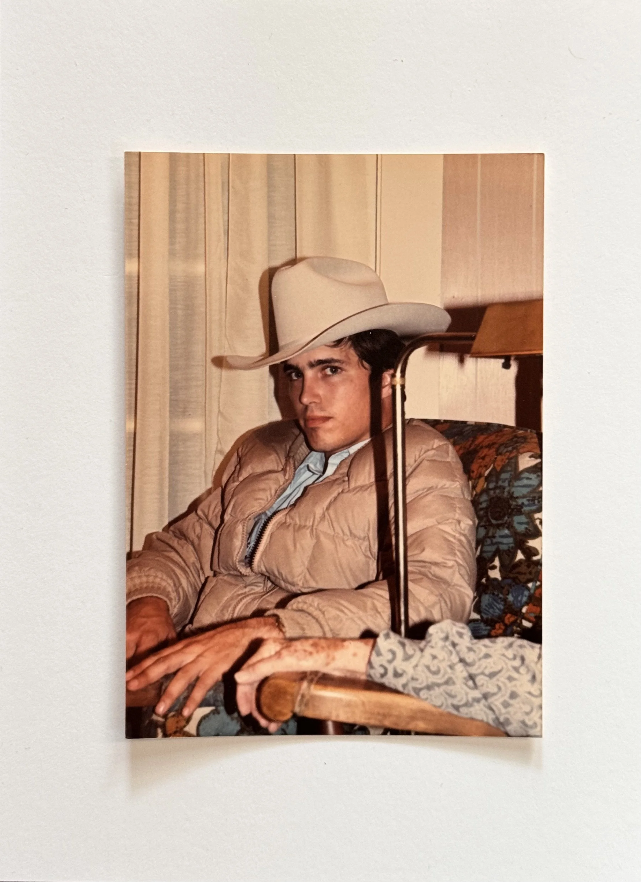 Young man sitting in a chair, wearing a tan puffer jacket, a wide-brimmed beige cowboy hat, and a light blue shirt, with a patterned cushion behind him.