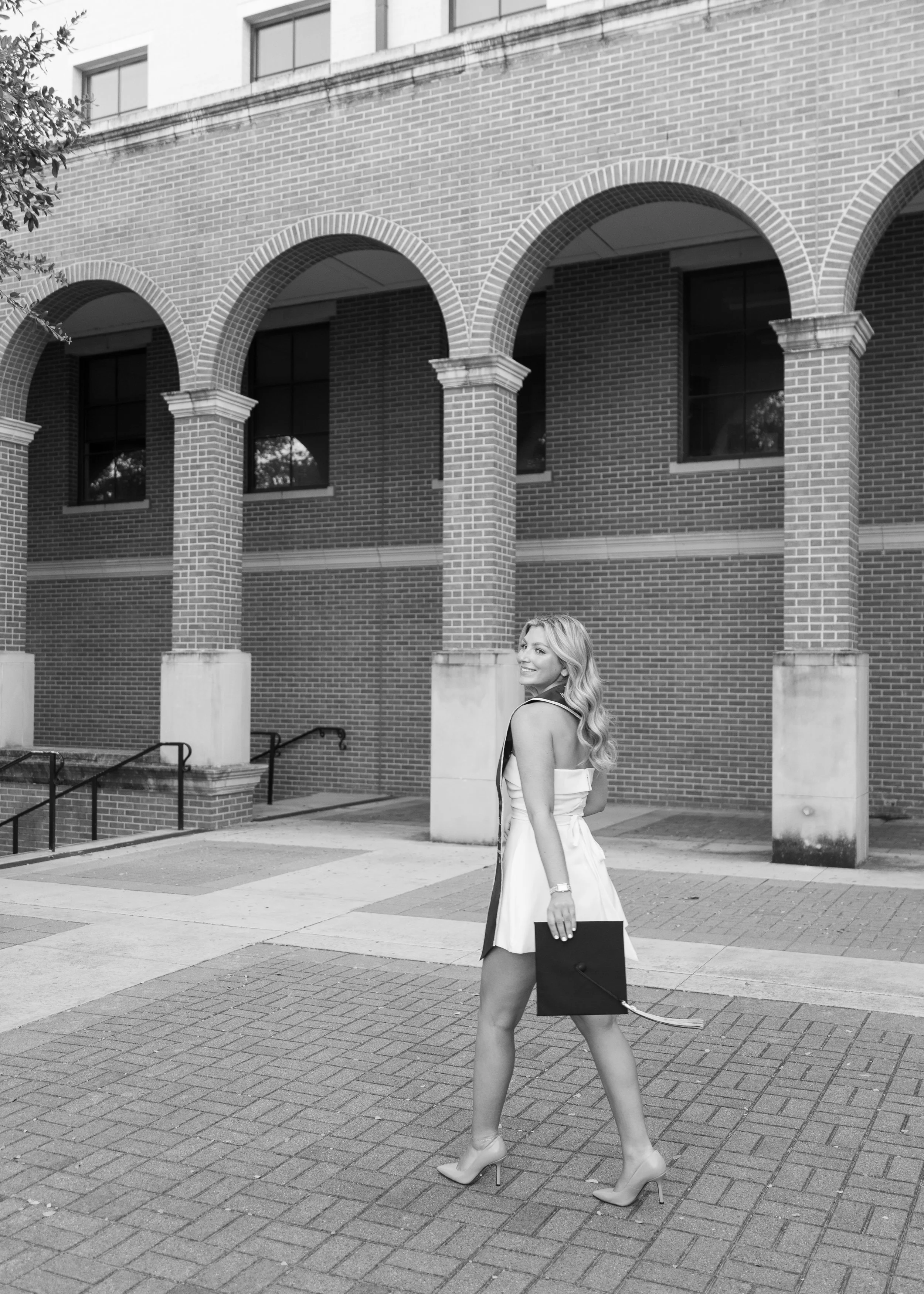 A young woman in a sleeveless dress with high heels, holding a graduation cap, walking outdoors in front of a brick building with arches, smiling and looking back.