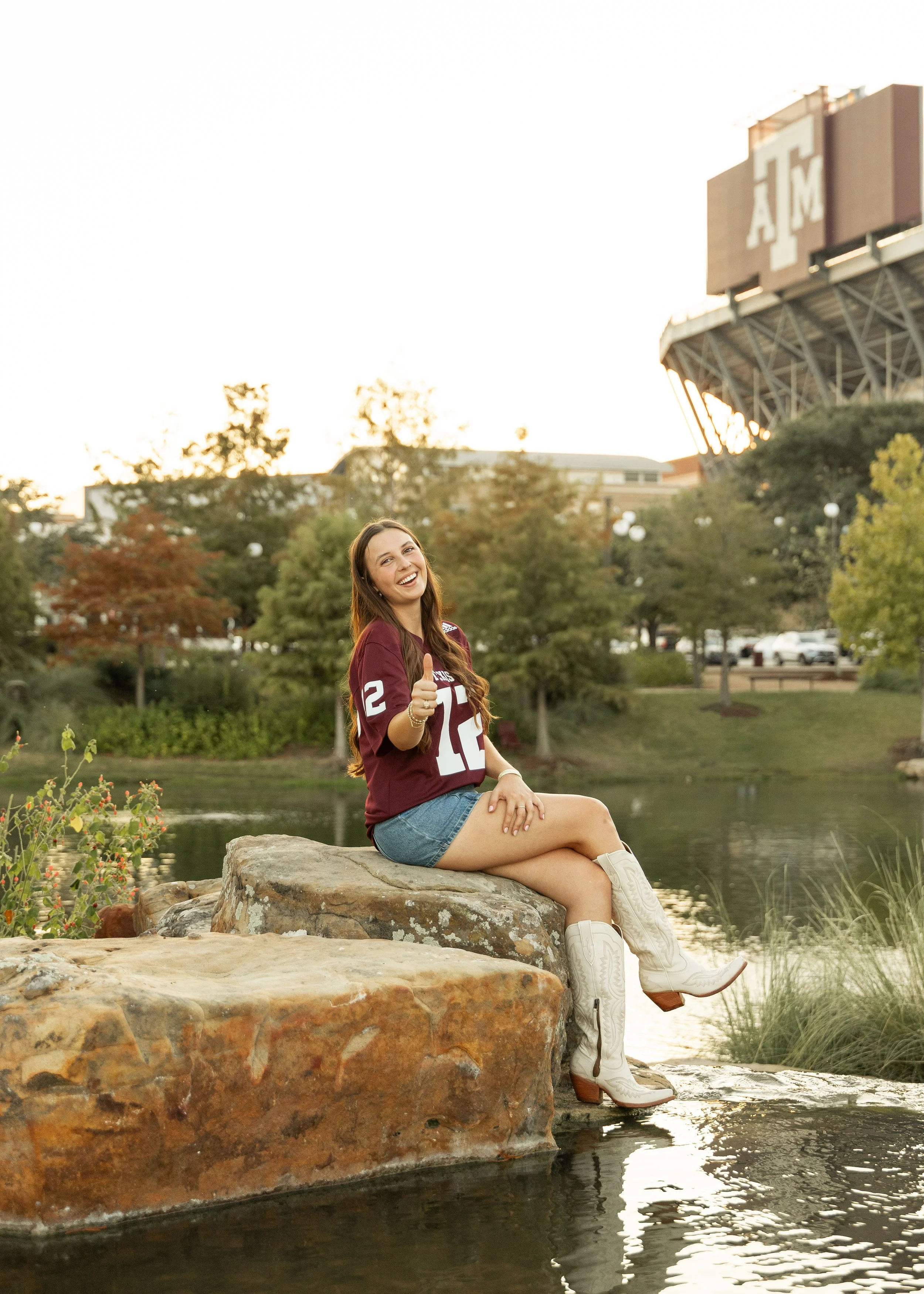 Young woman sitting on a large rock by a pond, wearing a maroon football jersey with the number 12, denim shorts, and white cowboy boots, giving a thumbs-up with a smile. In the background, there are trees, a campus building, and an athletic stadium 