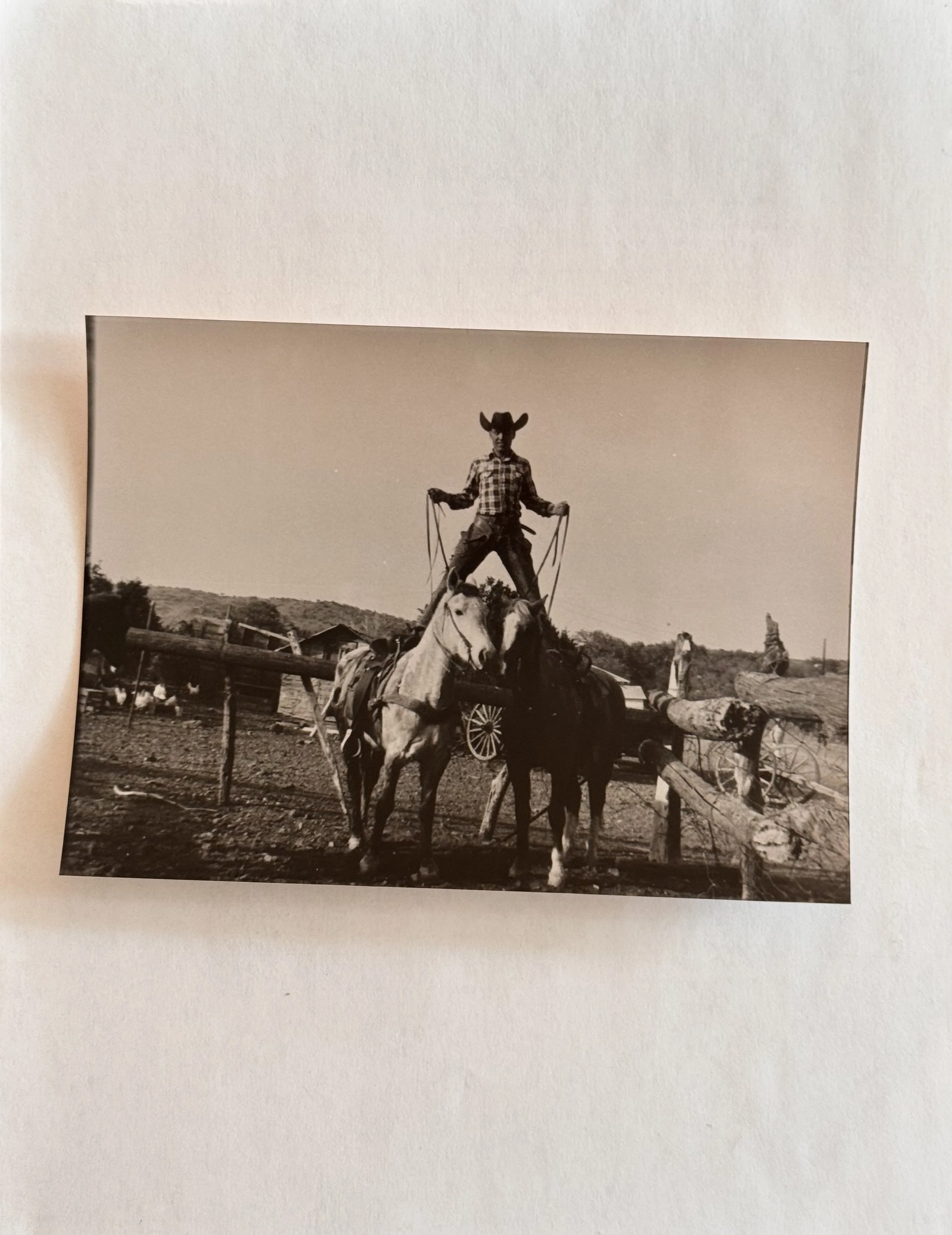 A black and white photo of a person wearing a western shirt, standing on a horse-drawn wagon, holding reins, with a rural landscape in the background.