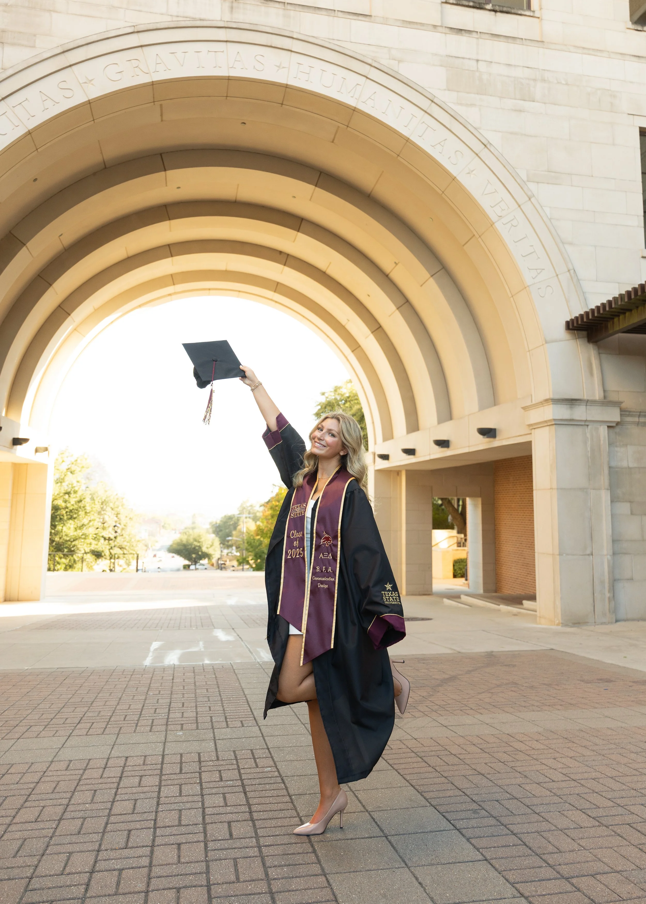 A young woman in graduation regalia standing under an archway, smiling, holding her cap in the air.