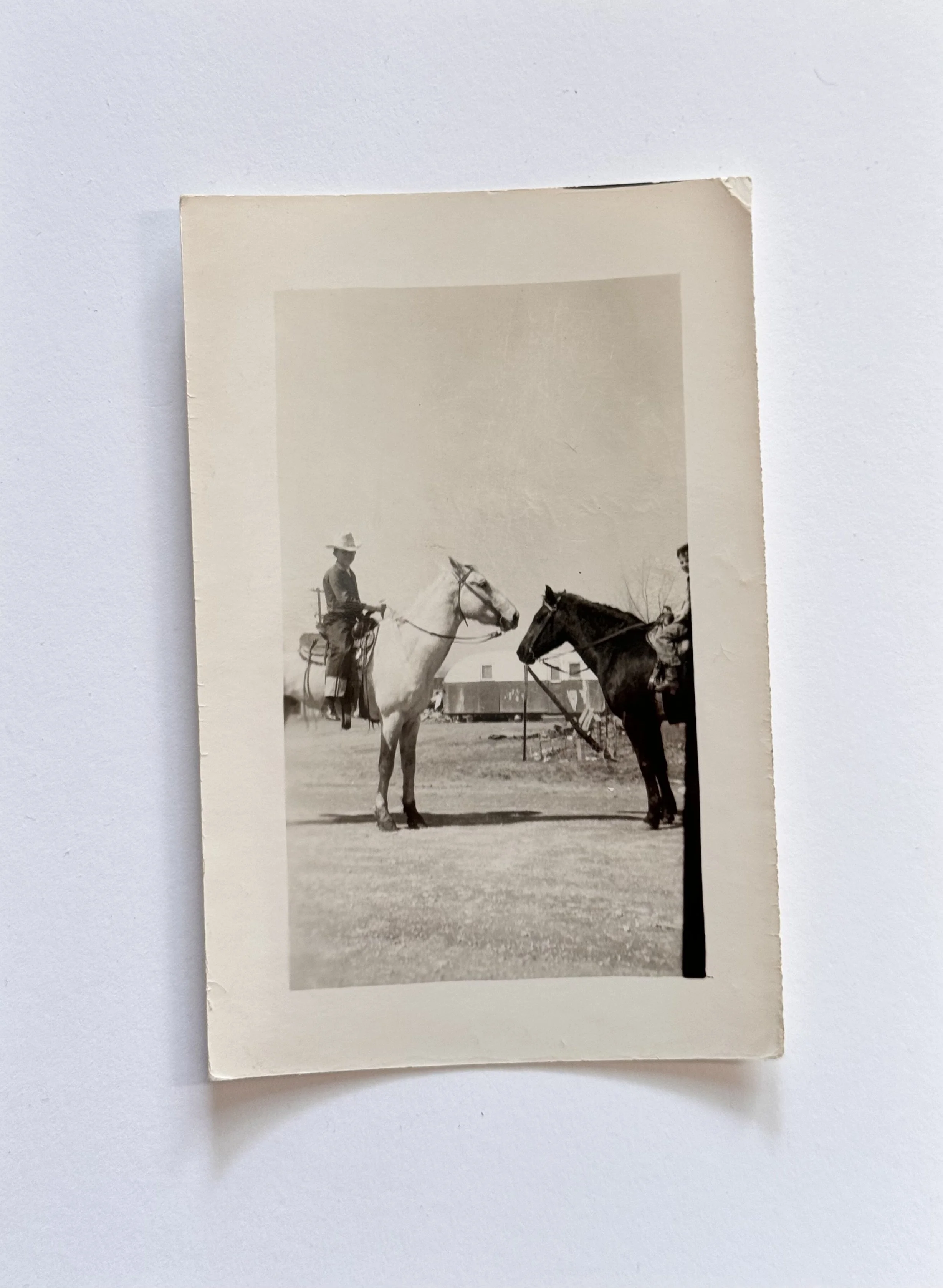 A black-and-white photo of two men on horseback, with one horse standing and the other possibly mounted, in an open field near a house or barn in the background.