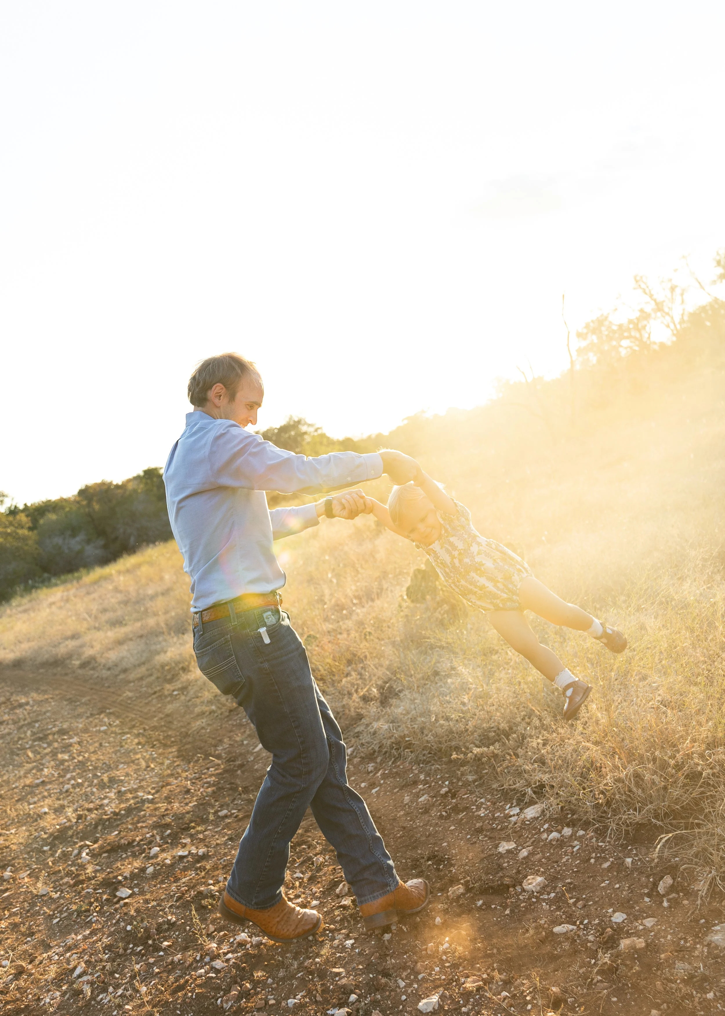 A man and a young girl are outside on a hill at sunset, playing. The man is spinning the girl around by holding her hands as she leans back in joyful motion.