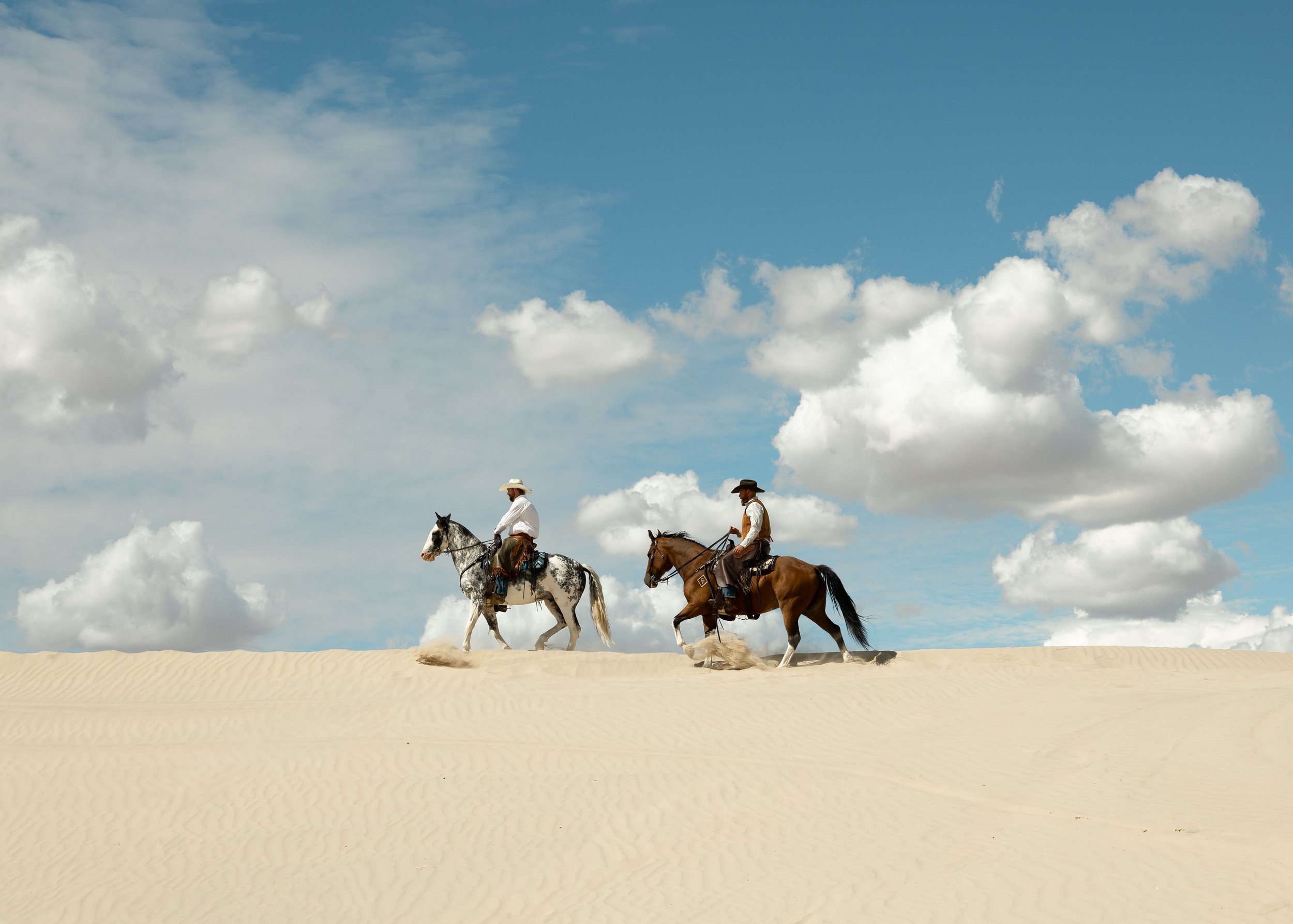 Two cowboys riding horses across sandy desert dunes under a partly cloudy sky.