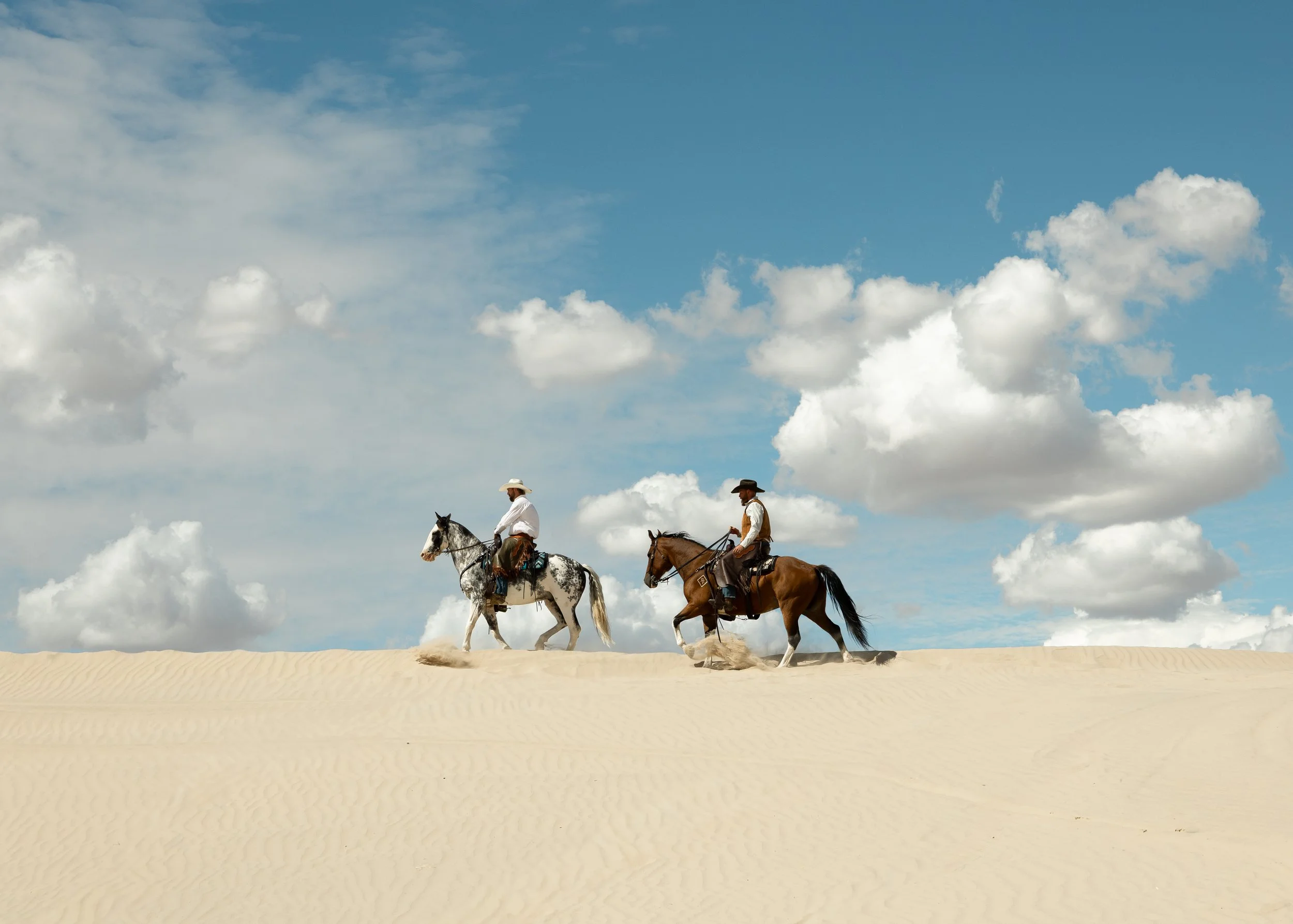 Two cowboys riding horses across a sandy desert under a blue sky with white clouds.