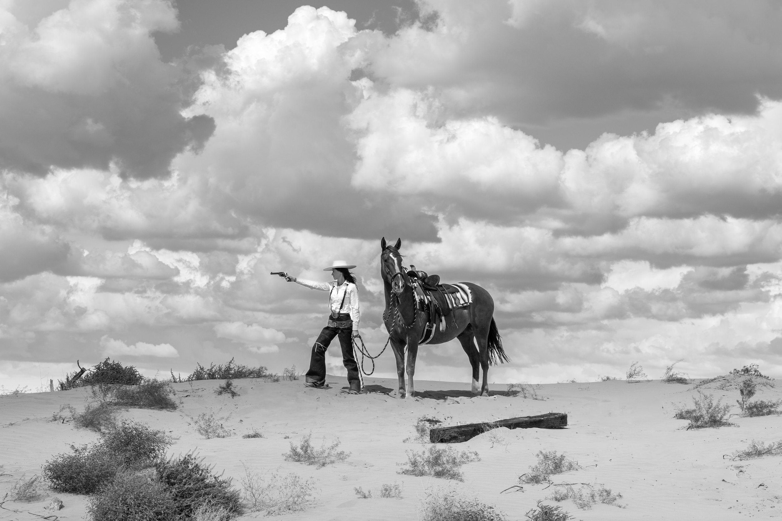 A person wearing a hat and western clothing, holding a gun in their right hand, stands next to a saddled horse in a desert landscape with sand, sparse bushes, and a cloudy sky.
