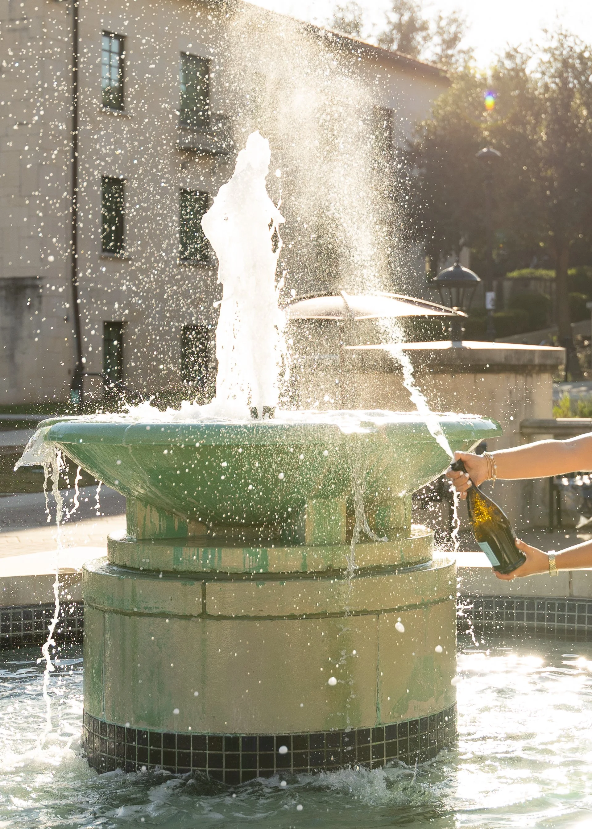 A decorative fountain with water spraying from the top, held by a person's hand with a bottle, in front of a building with windows, during daylight.