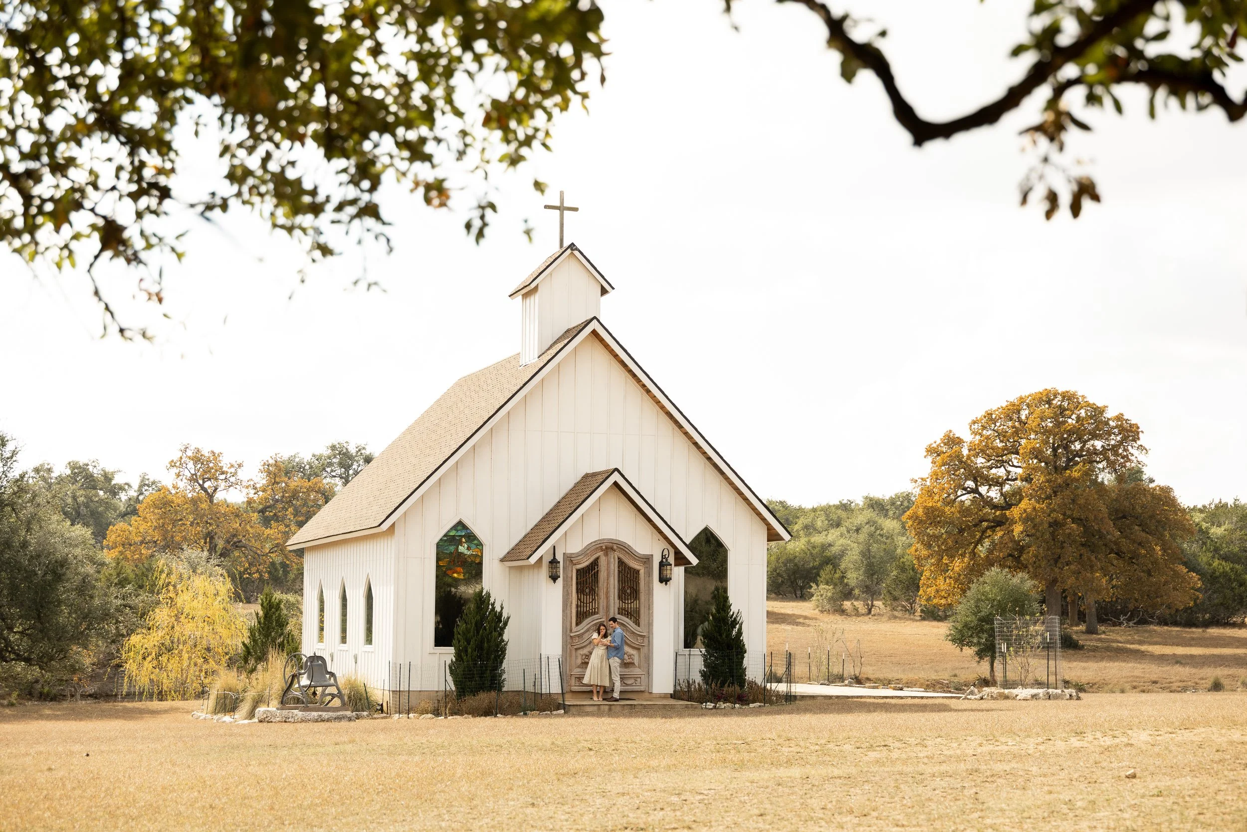 A small white church with a steeple and cross on top, surrounded by trees with autumn foliage, with a couple standing in front of the church entrance.
