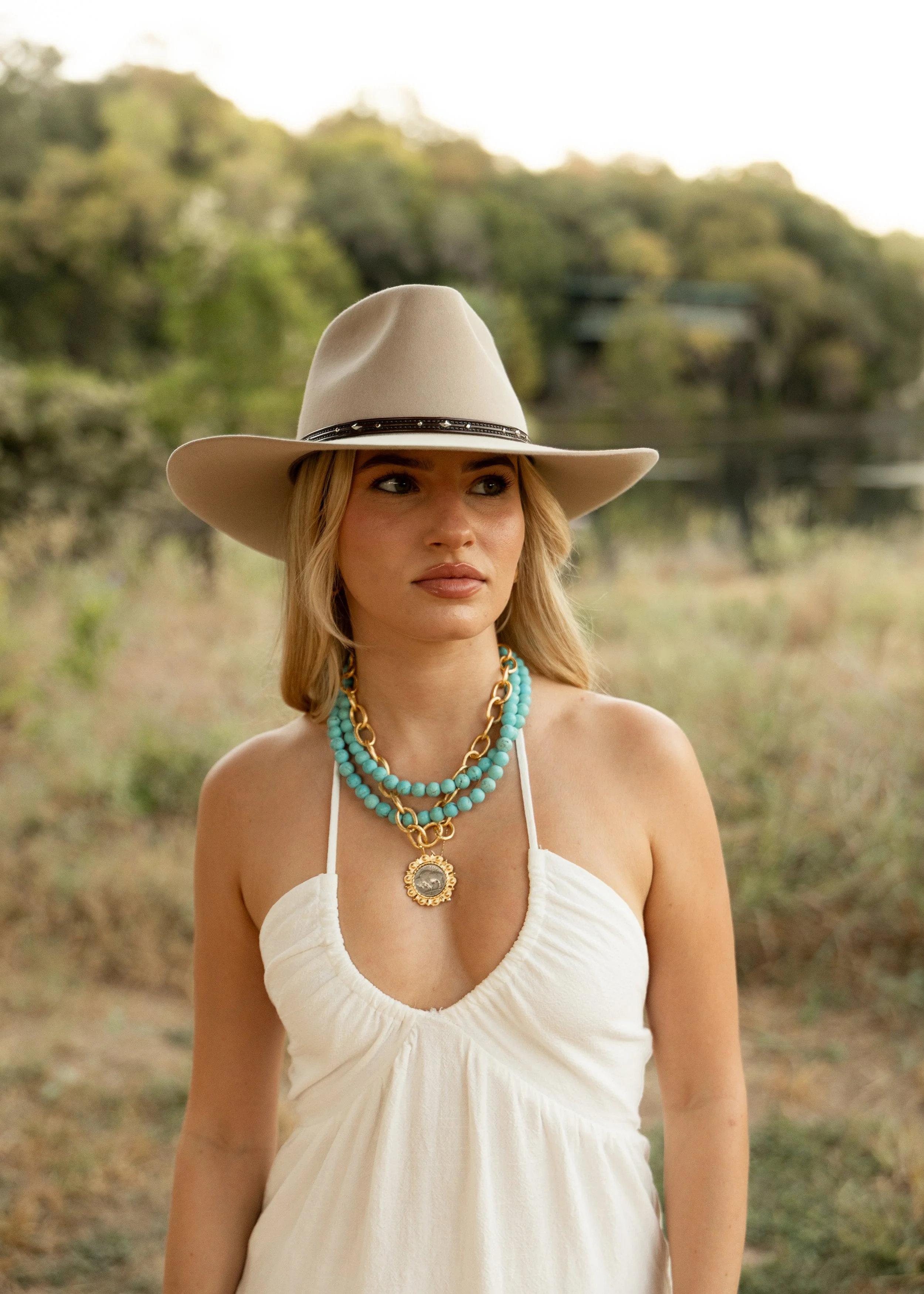 A woman wearing a beige wide-brimmed hat, layered turquoise and gold necklaces, and a white sleeveless dress, standing outdoors near a body of water with trees in the background.
