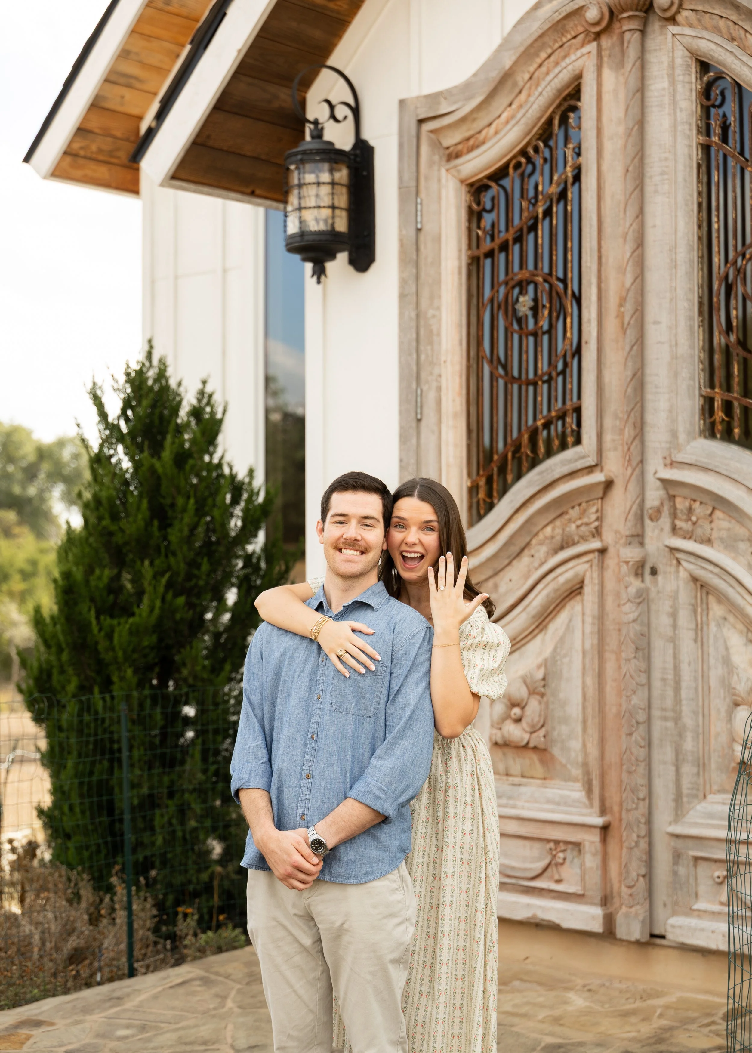 A smiling couple standing outside a house, with the woman showing off a ring on her finger, suggesting an engagement, near a decorative wooden door.