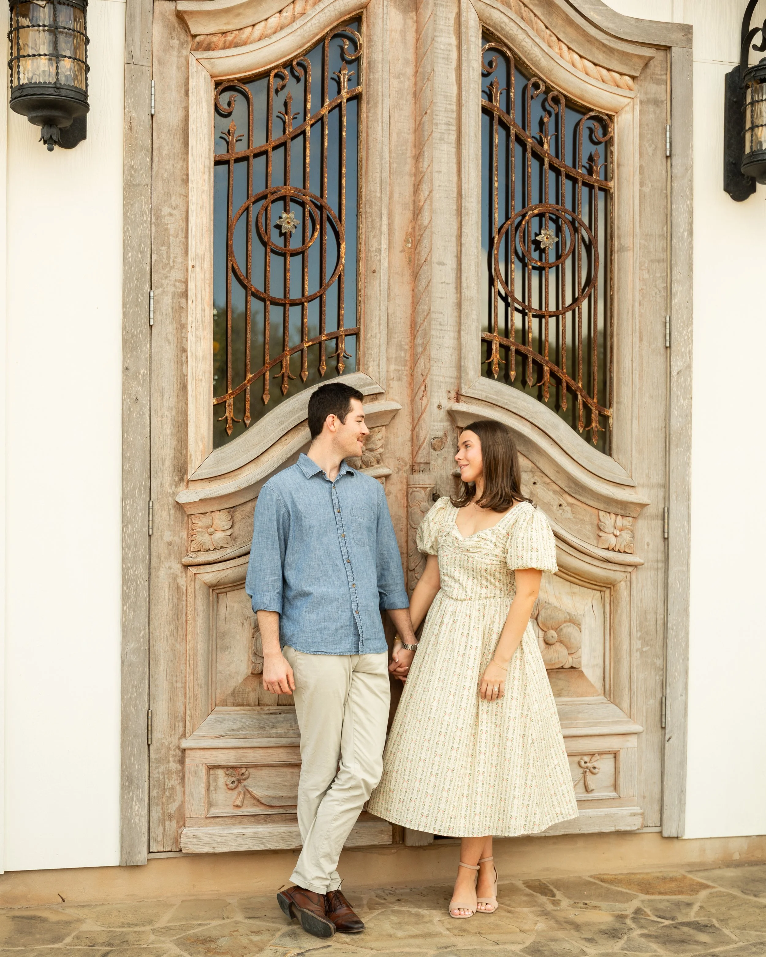 A young couple holding hands and smiling at each other standing in front of a large rustic wooden door with ornate ironwork.
