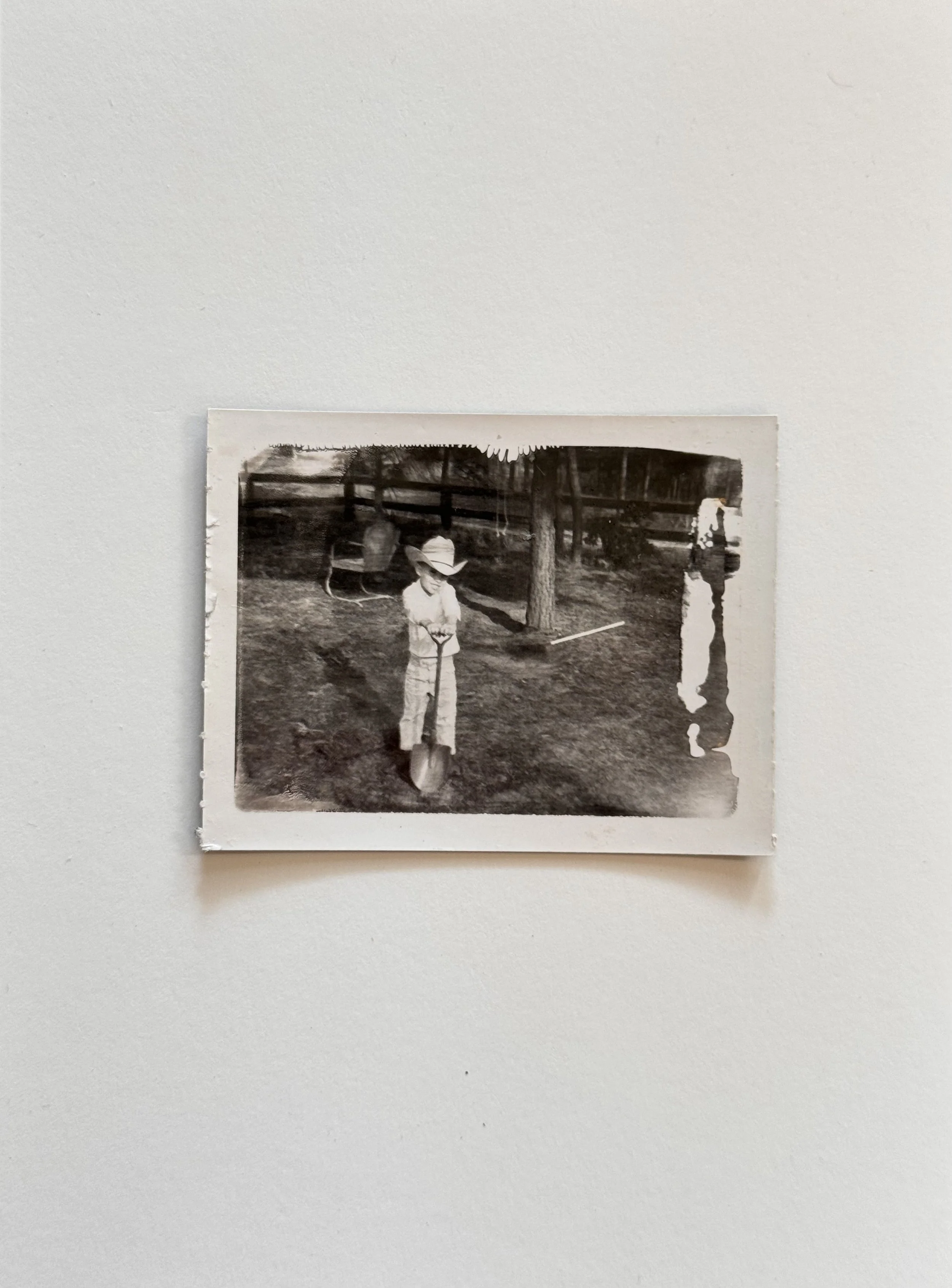 A black and white photograph of a young boy wearing a cowboy hat, holding a shovel, standing outdoors near a tree and a fence.
