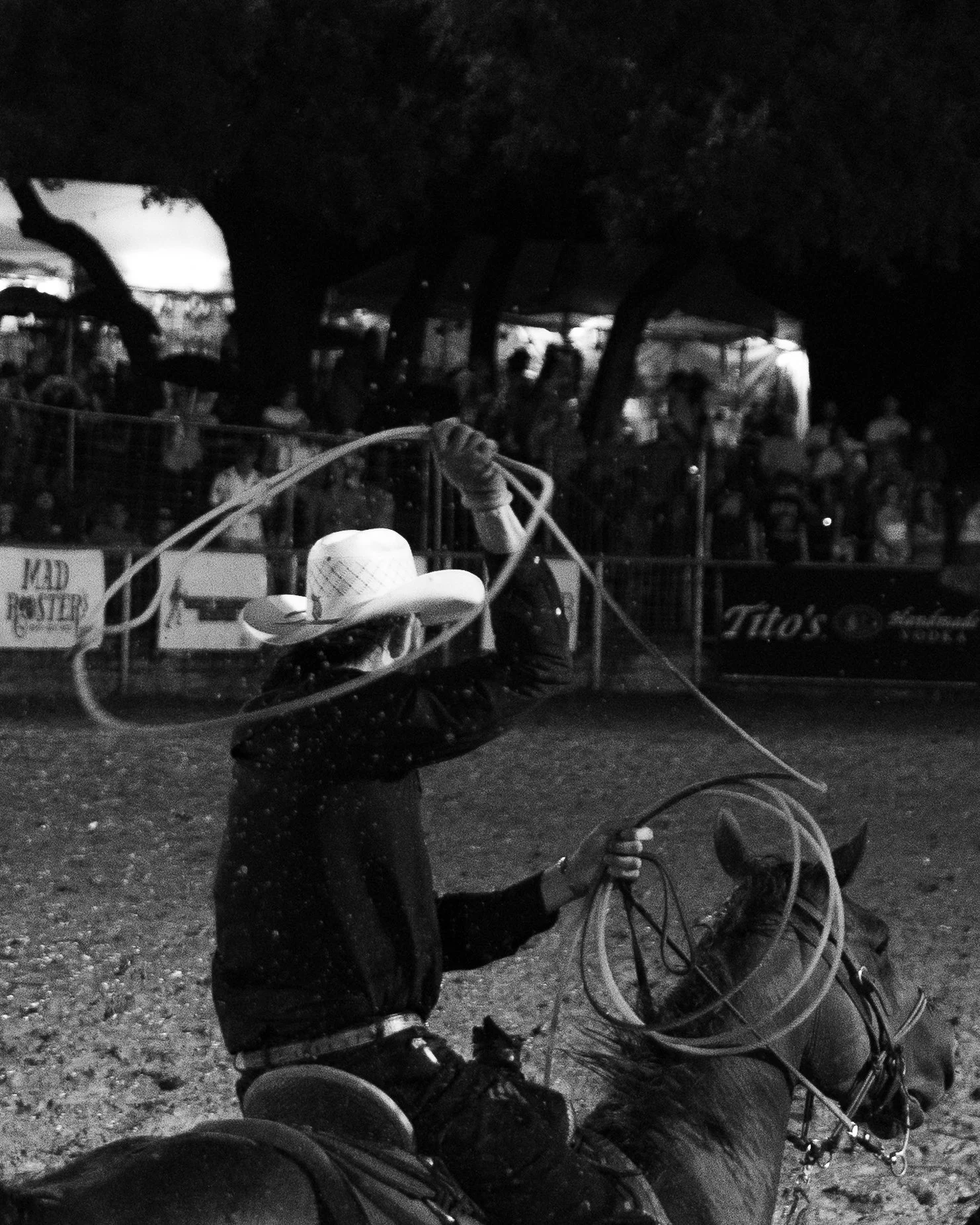 A person wearing a cowboy hat and dark clothing is riding a horse at a rodeo or similar event, holding a lasso against a crowd of spectators under trees.