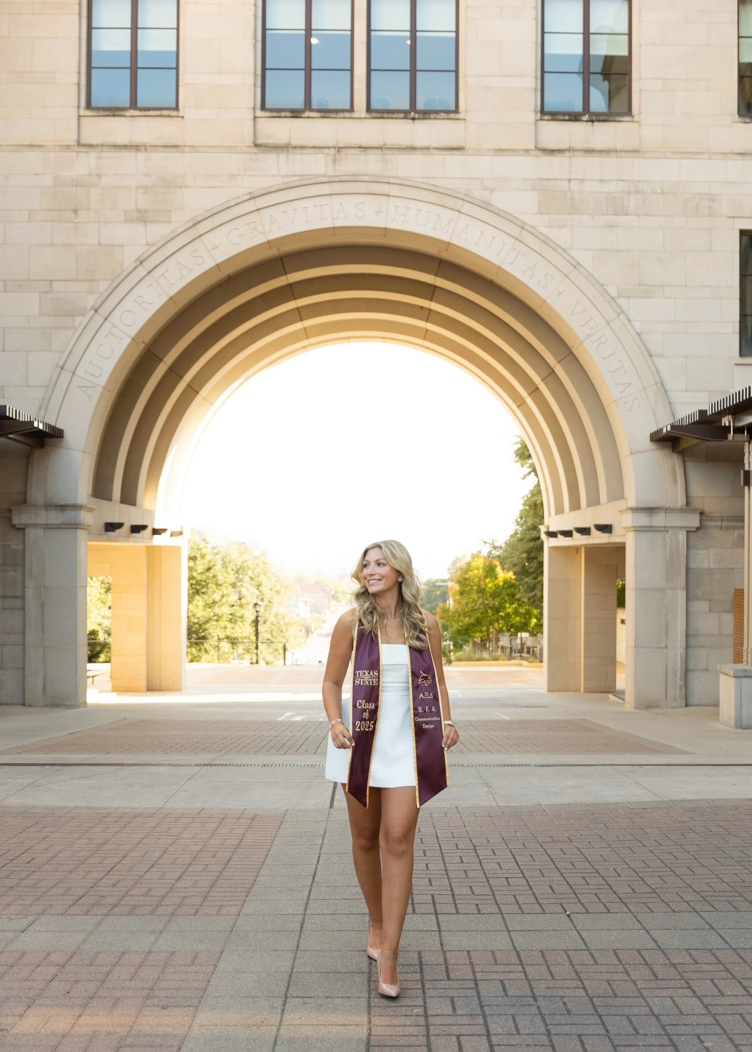 Graduation cap and gown woman standing under archway on university campus with sunlight behind