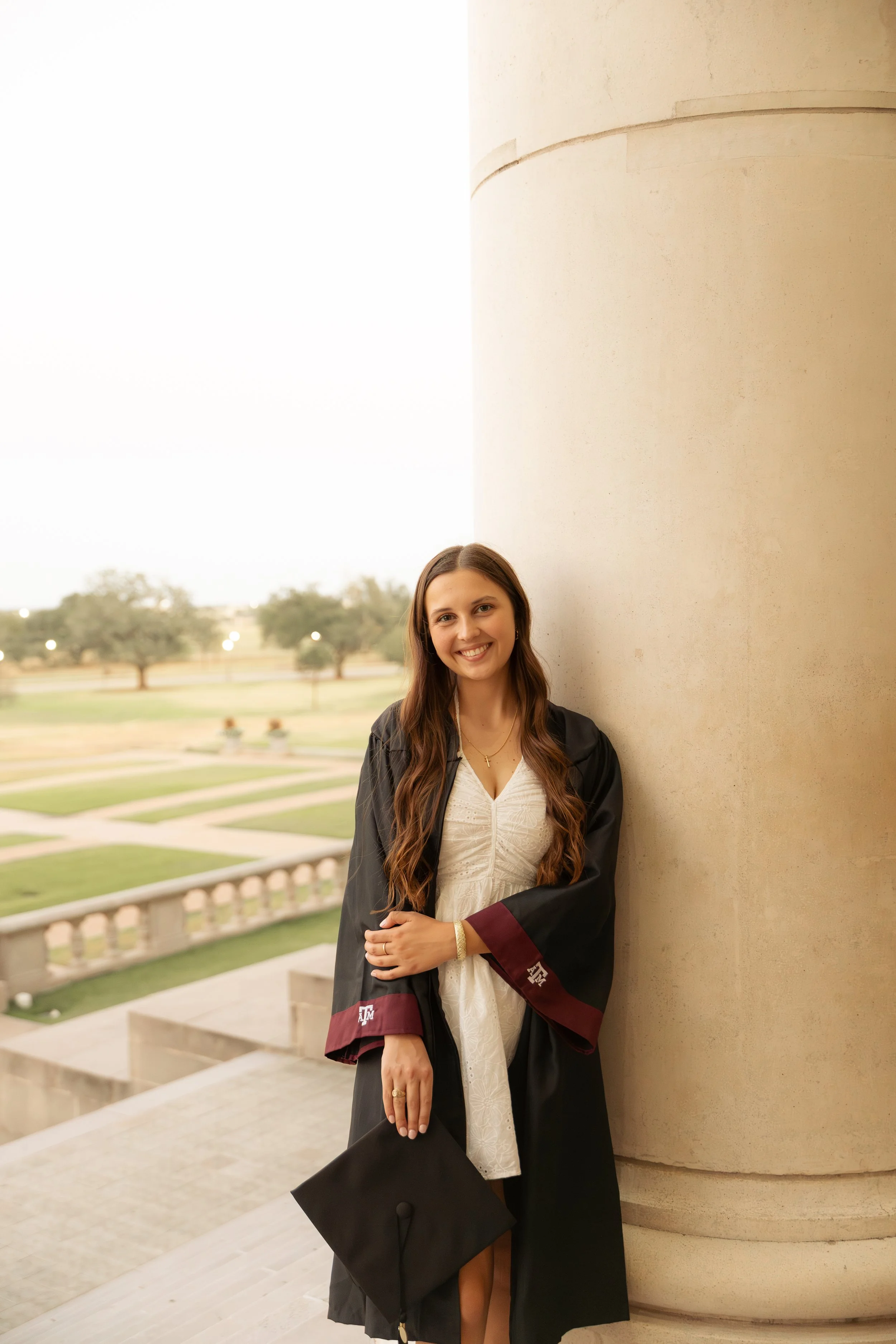 A young woman in graduation attire standing outdoors by a large column, holding a graduation cap, and smiling.