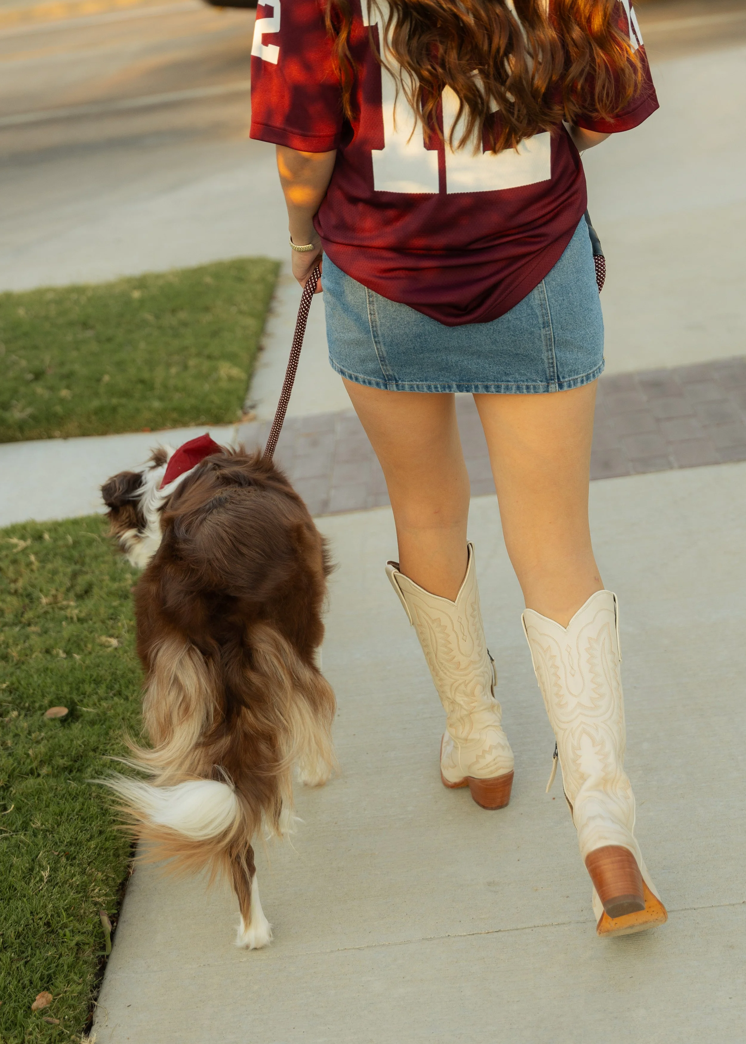 A woman walking a dog on a sidewalk. The woman is wearing a football jersey, a denim skirt, and cowboy boots. The dog is a fluffy breed with a red vest and a Santa hat on. The scene suggests a casual outdoor walk.