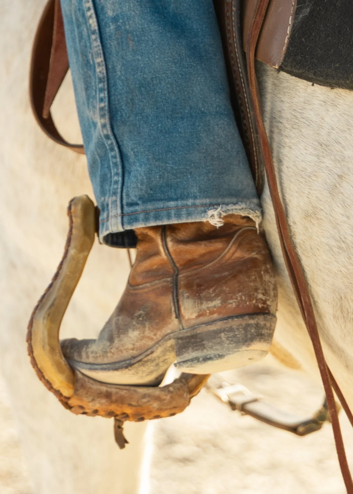 Close-up of a cowboy boot with a worn leather finish and a heel, sitting next to a saddle on a light-colored surface.