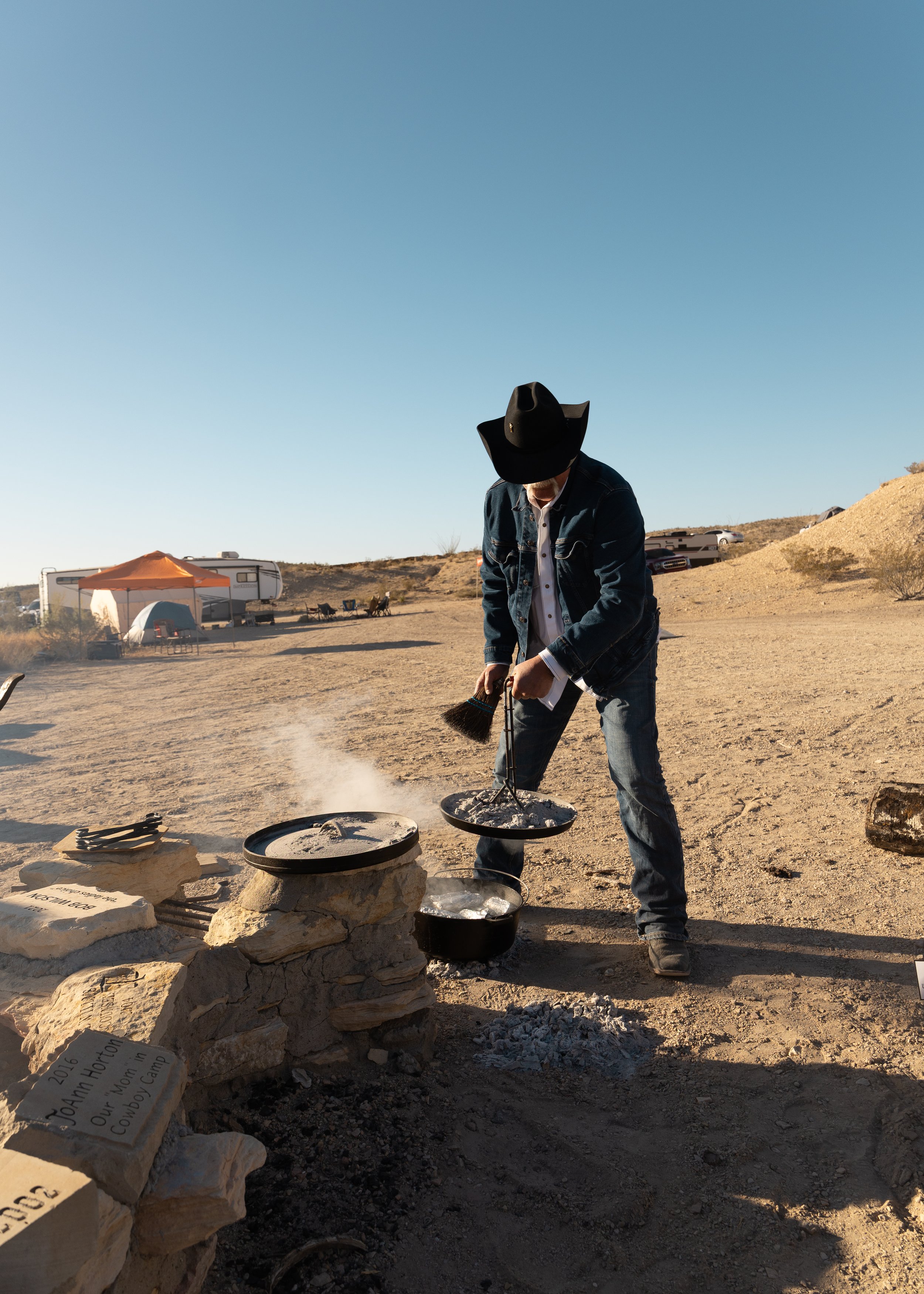 A person in a cowboy hat and denim jacket cooking over an outdoor campfire in a desert landscape with camping trailers and tents in the background.