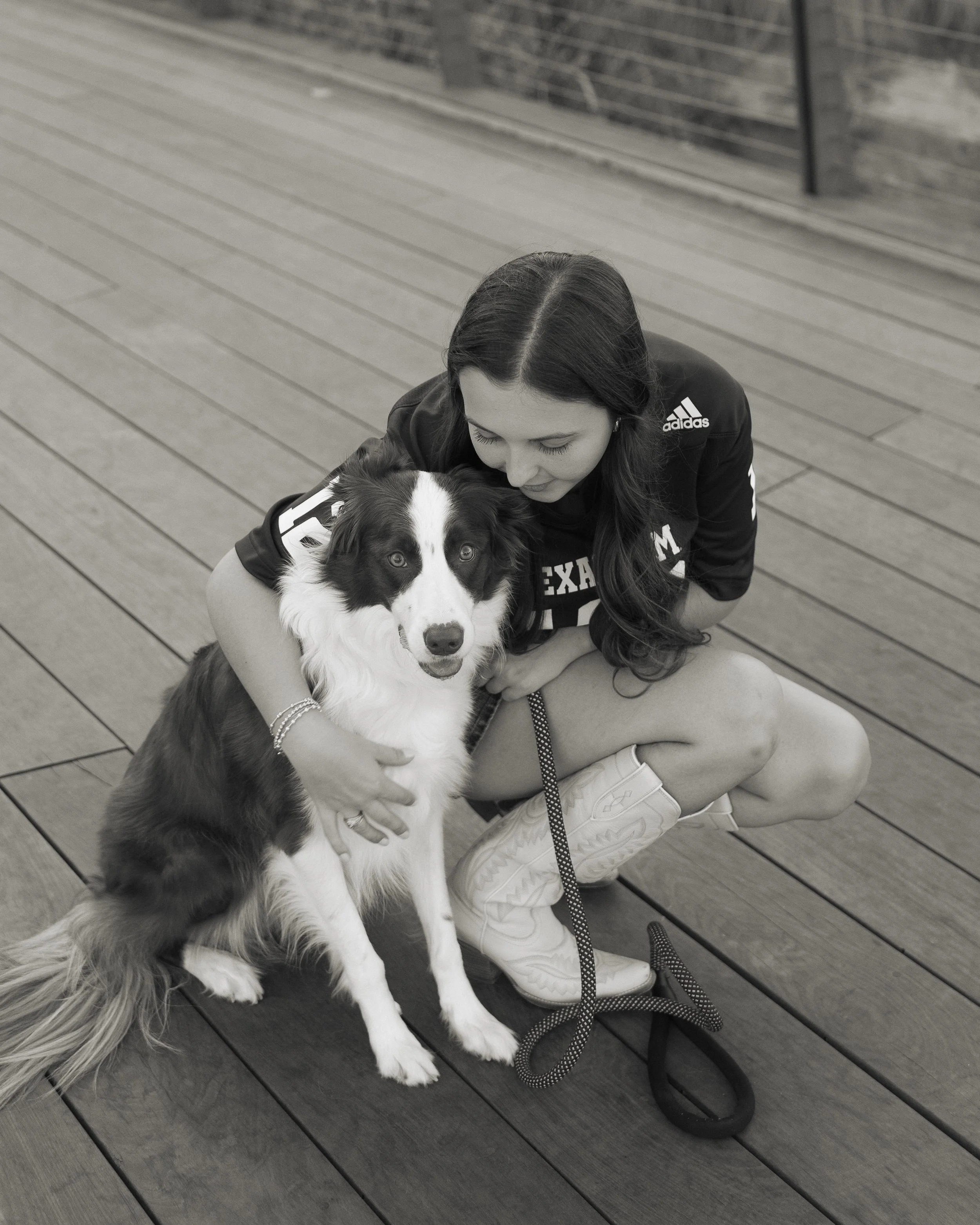 A woman wearing a sports jersey and cowboy boots kneels on a wooden deck, hugging a black and white Border Collie dog. The woman is looking down at the dog, which is facing forward and sitting on the deck.
