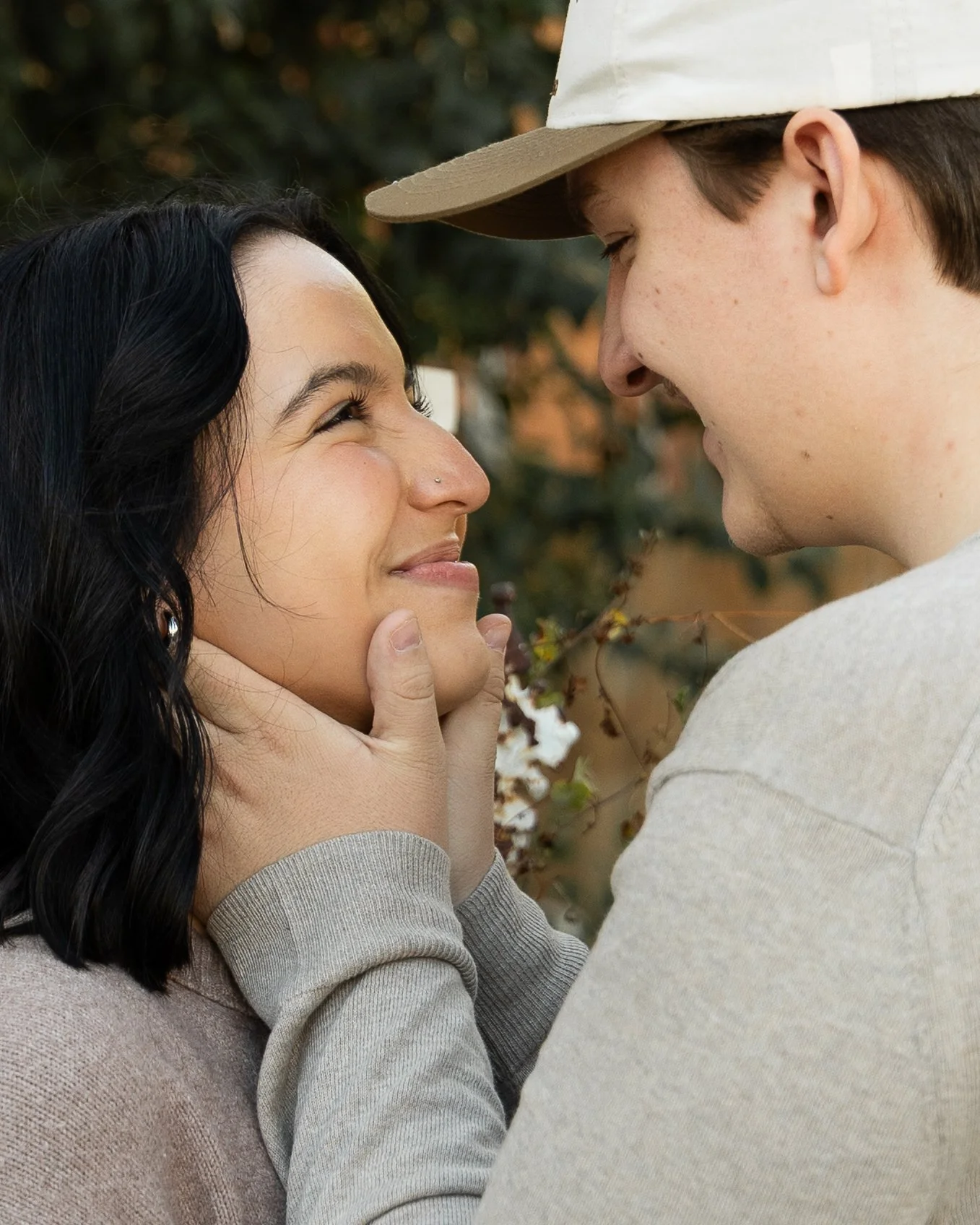 A young couple is close together outdoors, with the man gently holding the woman's face, both smiling and gazing into each other's eyes.