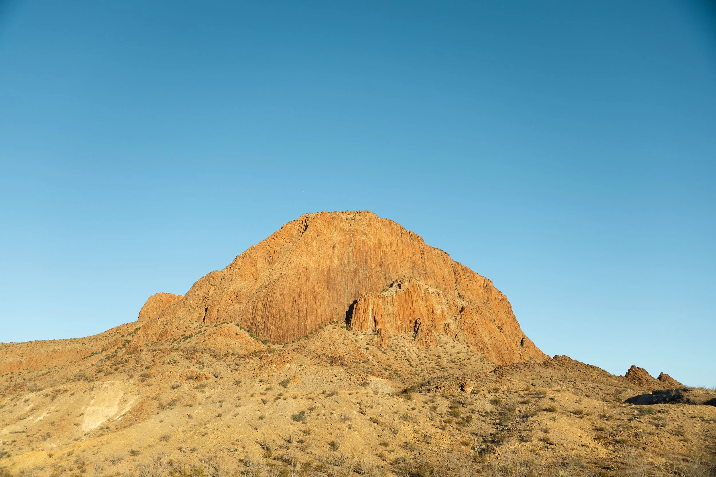 A large reddish-brown rock formation in a desert landscape beneath a clear blue sky.