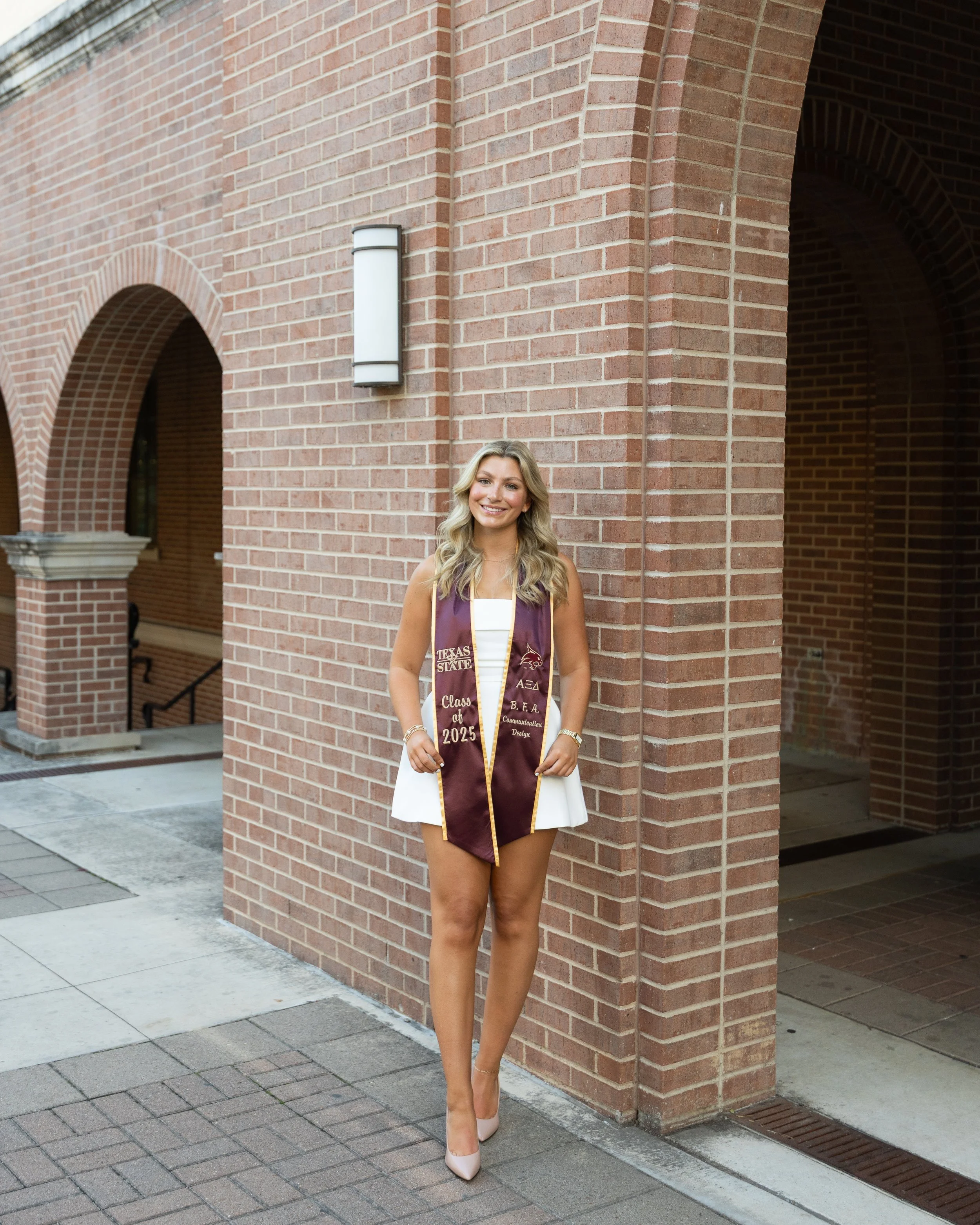 A young woman standing outside in front of a brick building, wearing a white dress, beige high heels, and a maroon graduation stole with gold trim and text.