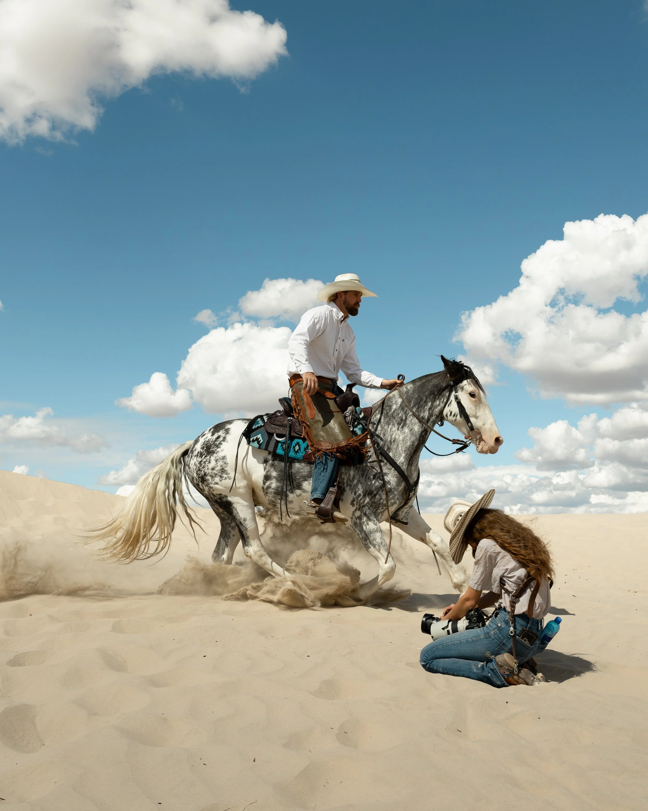A man riding a horse in a desert, while a woman kneeling on the sand takes a photo with a camera. The sky is blue with white clouds.