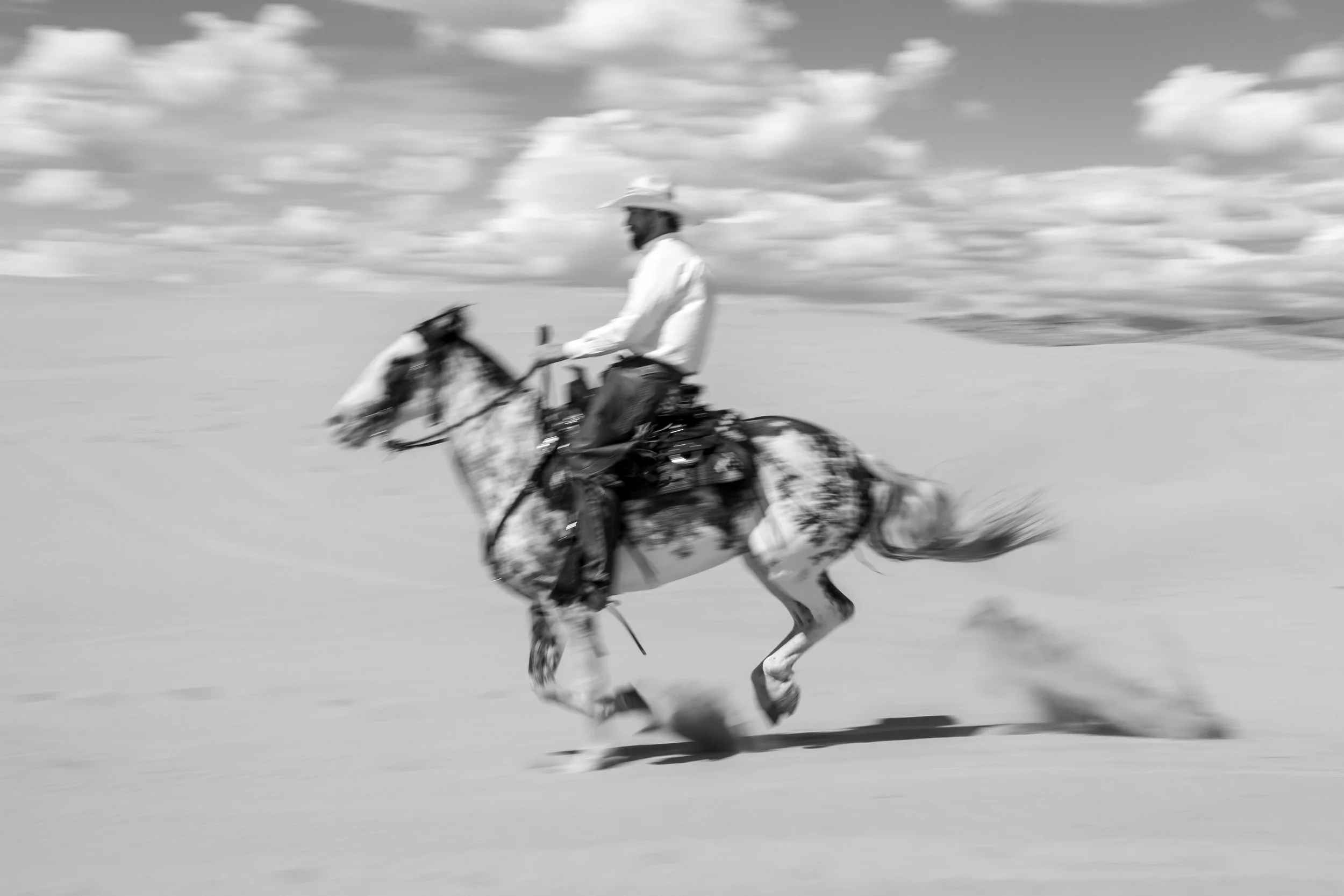 A man riding a horse at a fast pace in a desert landscape with clouds in the sky.