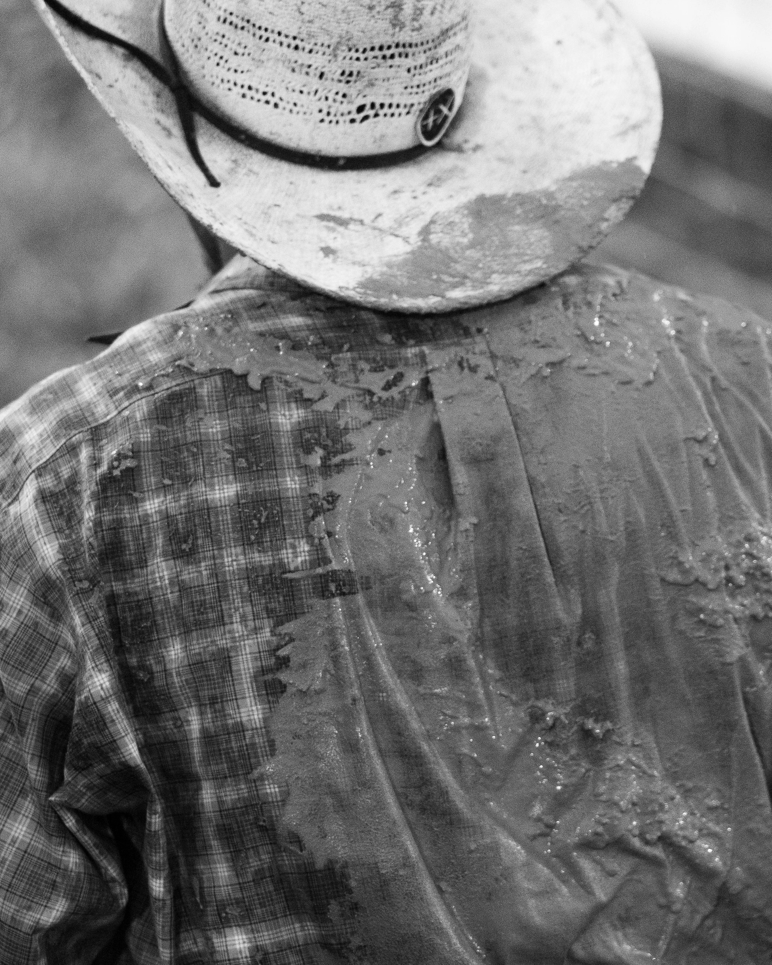 A person wearing a cowboy hat and plaid shirt, with wet, muddy clothing, seen from behind.