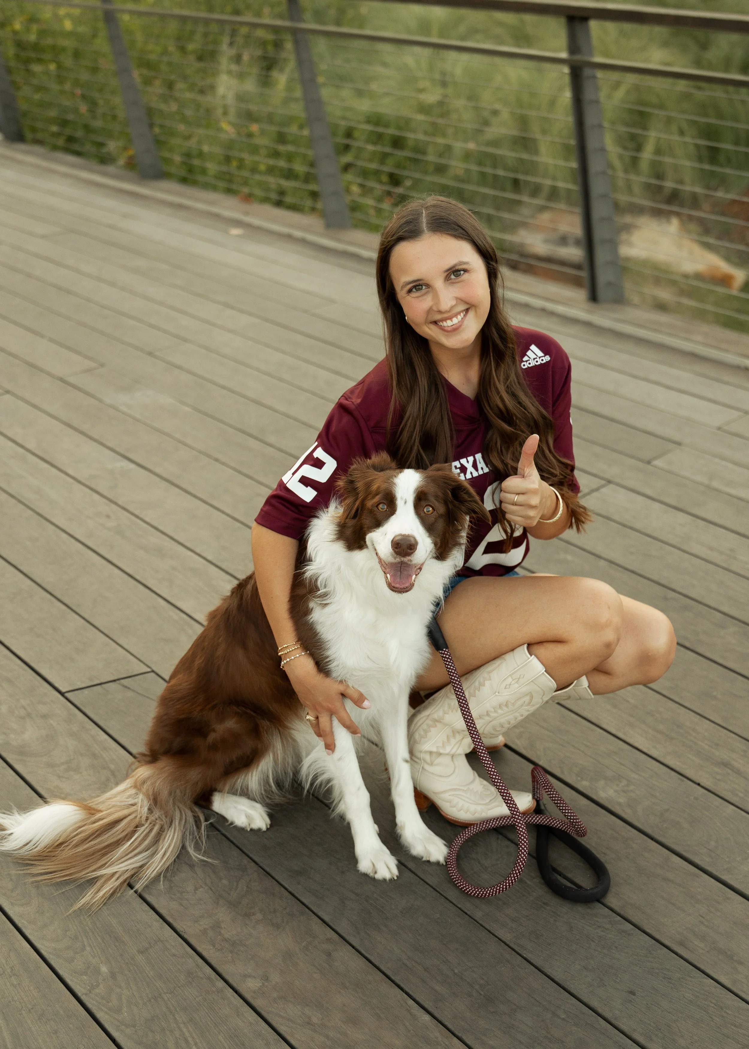A young woman with long brown hair kneeling on a wooden bridge, smiling and giving a thumbs-up, with her Border Collie sitting next to her.