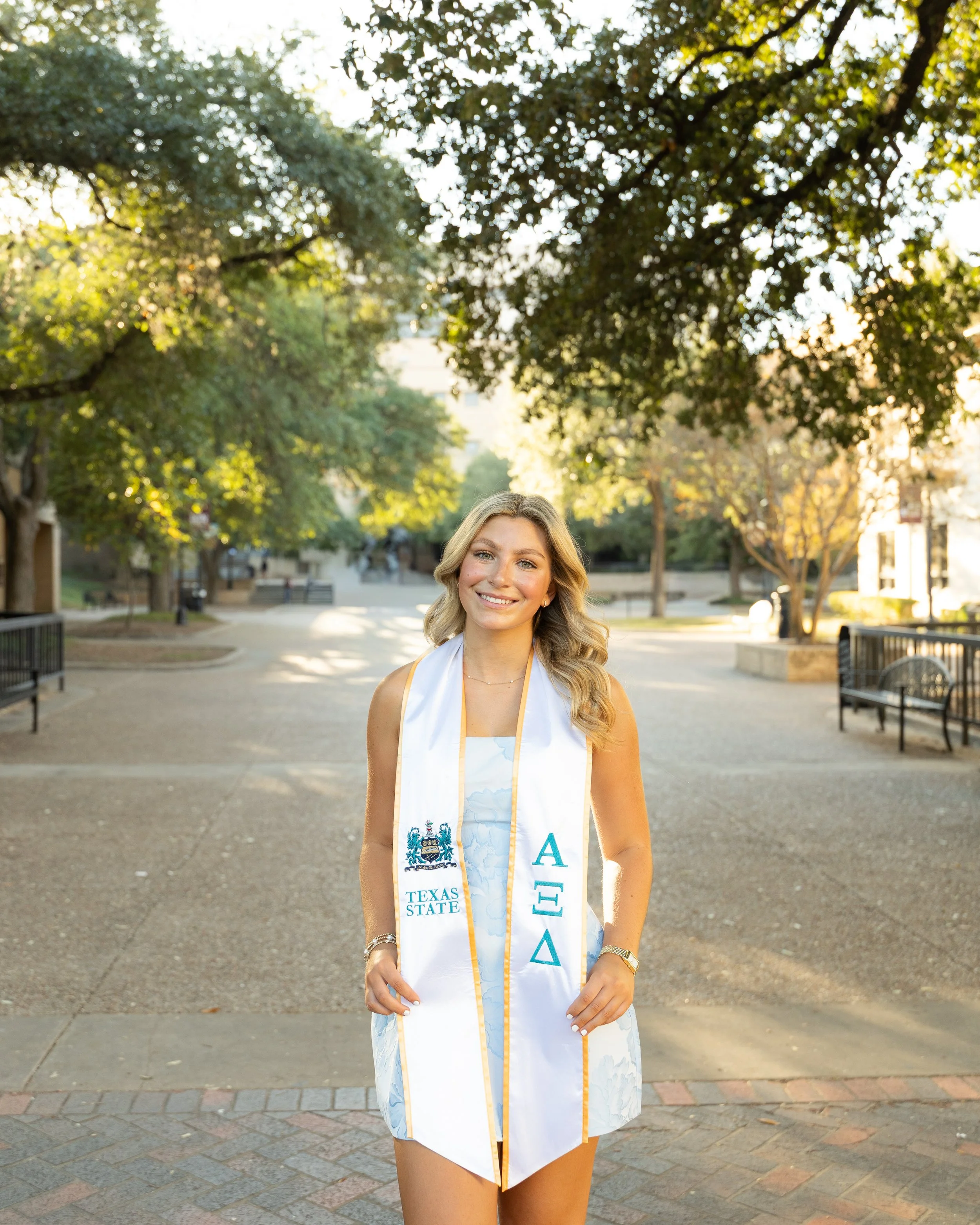 A young woman stands outdoors on a pathway, smiling, wearing graduation attire with a sash that has the words 'Texas State' and Greek letters, in between trees and benches on a college campus in the late afternoon.