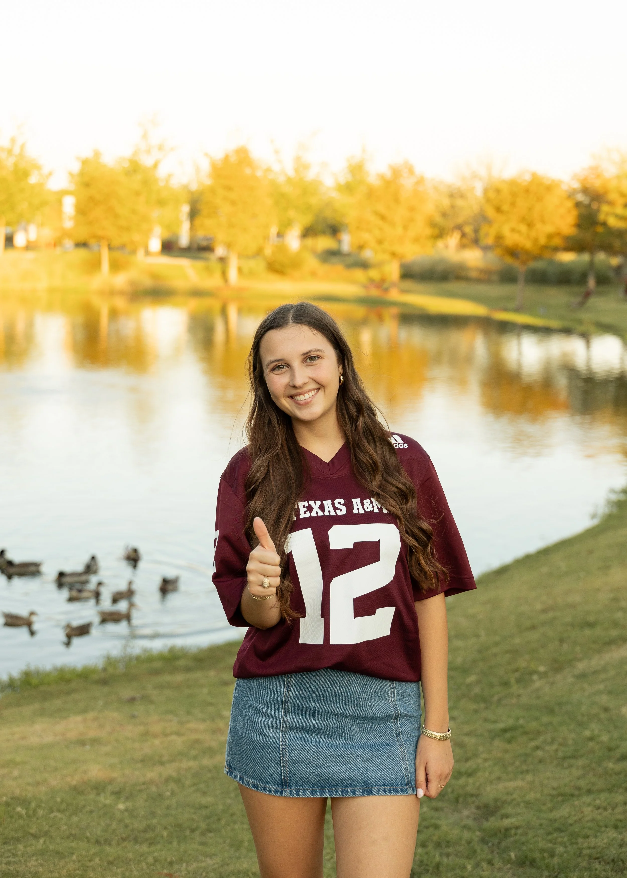 A young woman standing outdoors near a lake, wearing a maroon Texas A&M jersey with the number 12, smiling and giving a thumbs-up. Ducks are swimming in the lake, and trees with fall foliage are in the background.