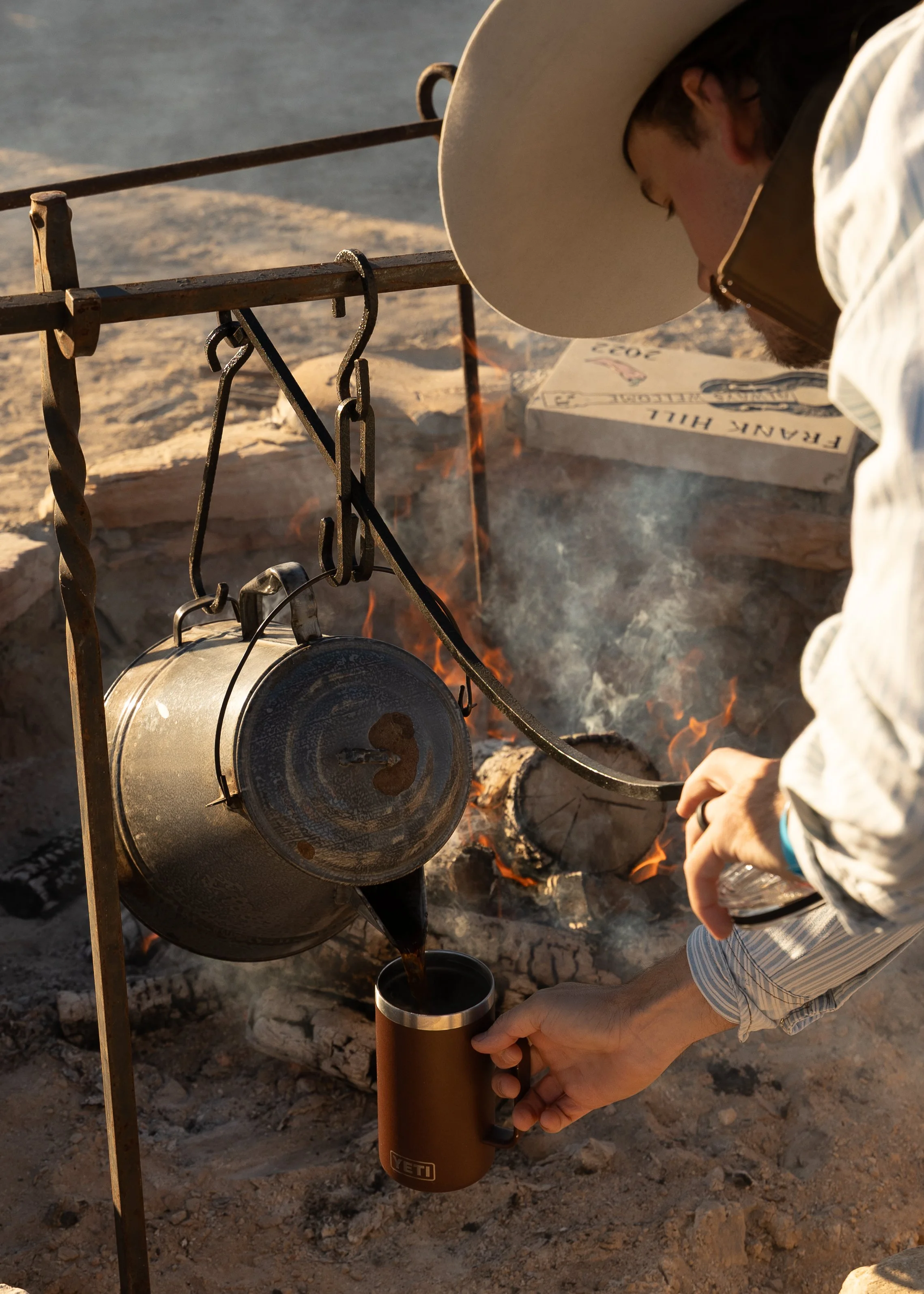 Person wearing a cowboy hat and light-colored jacket pouring coffee from a pot into a YETI mug over an open campfire.