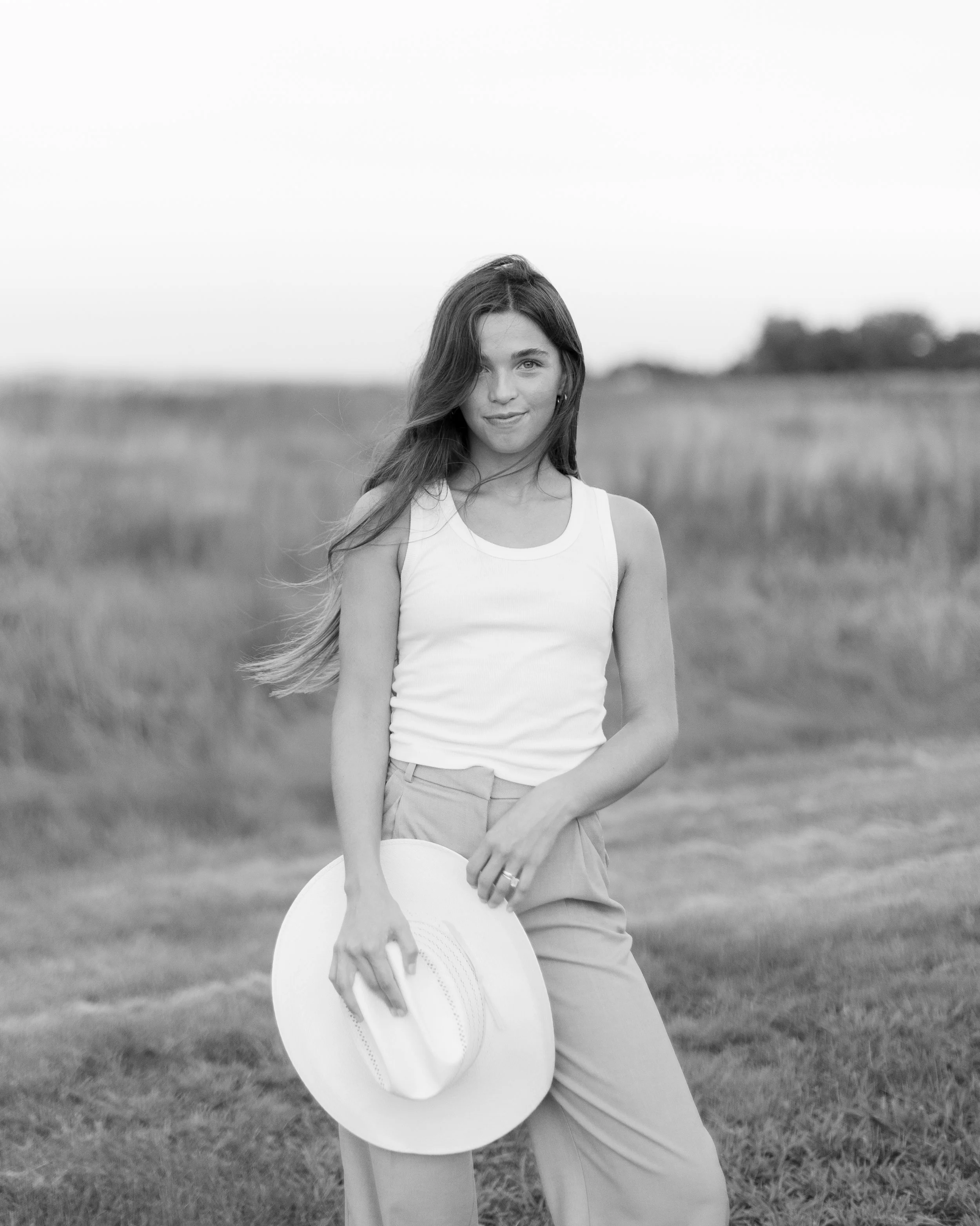A young woman stands in an open field, holding a wide-brimmed hat, wearing a tank top and high-waisted pants, with her hair flowing in the wind.