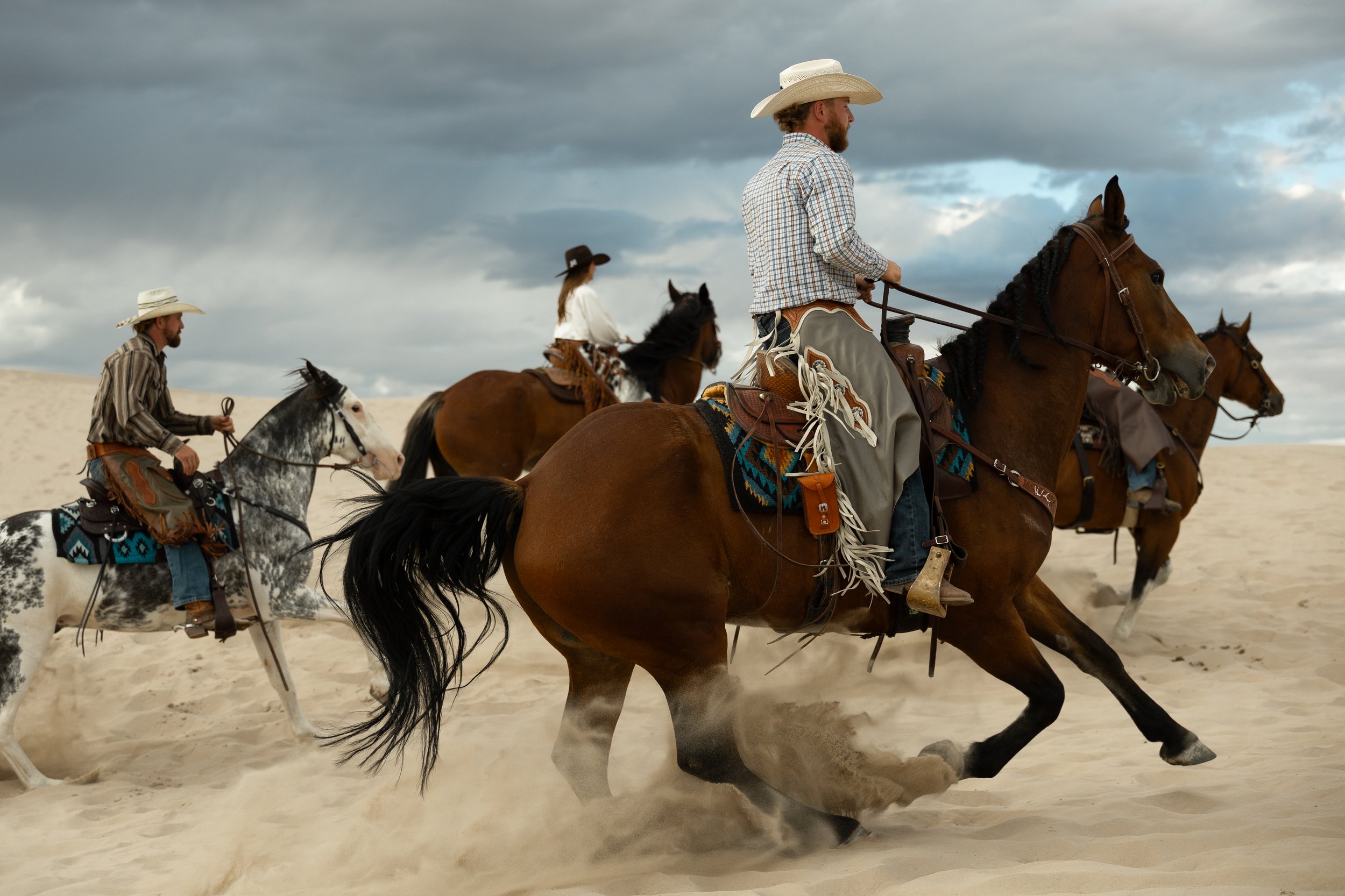 Group of four people riding horses across sandy desert with cloudy sky.