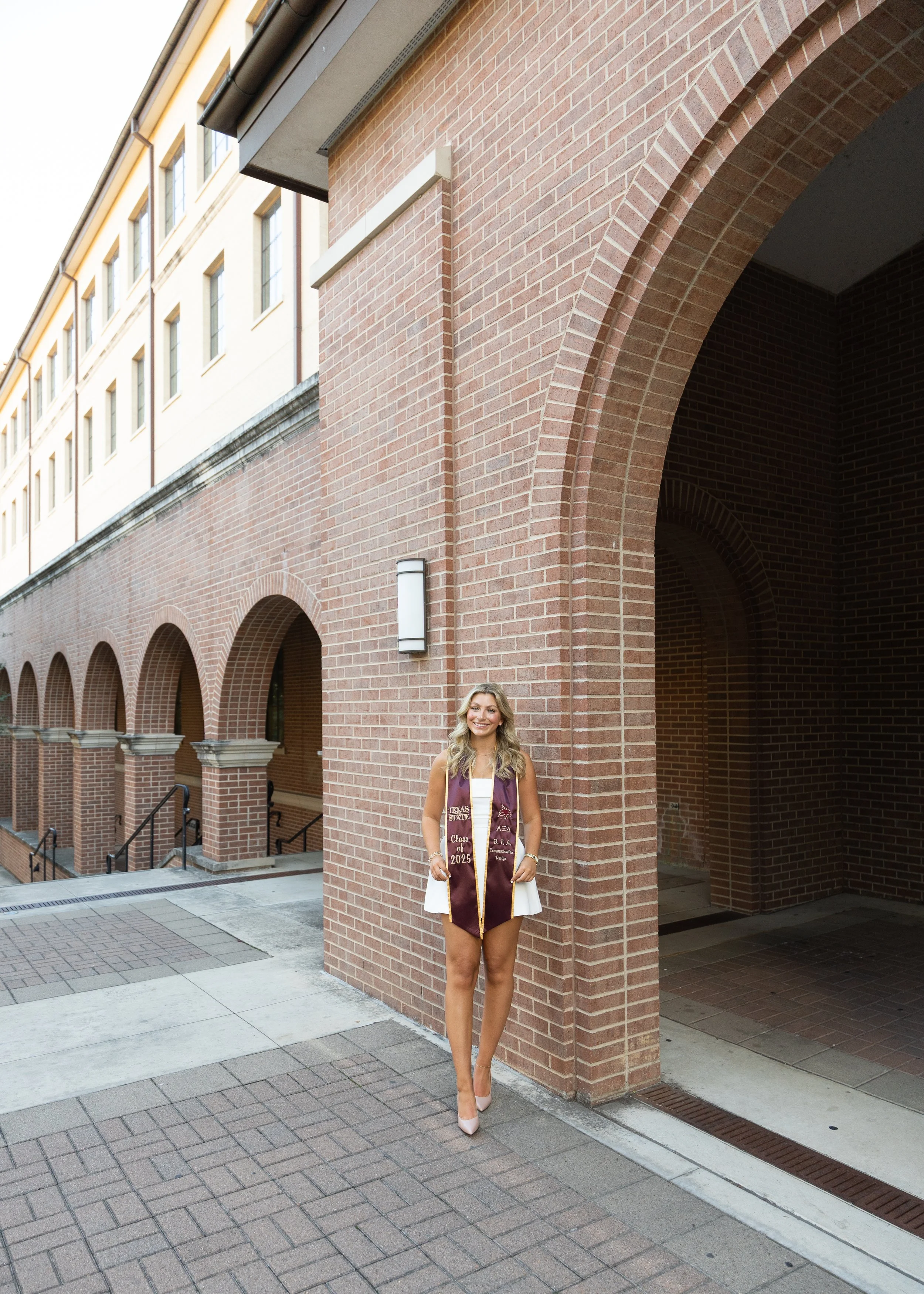 A young woman in a white dress and beige high heels stands outside on a sidewalk, wearing a maroon graduation stole with gold accents and text, in front of a brick building with arched entryways.