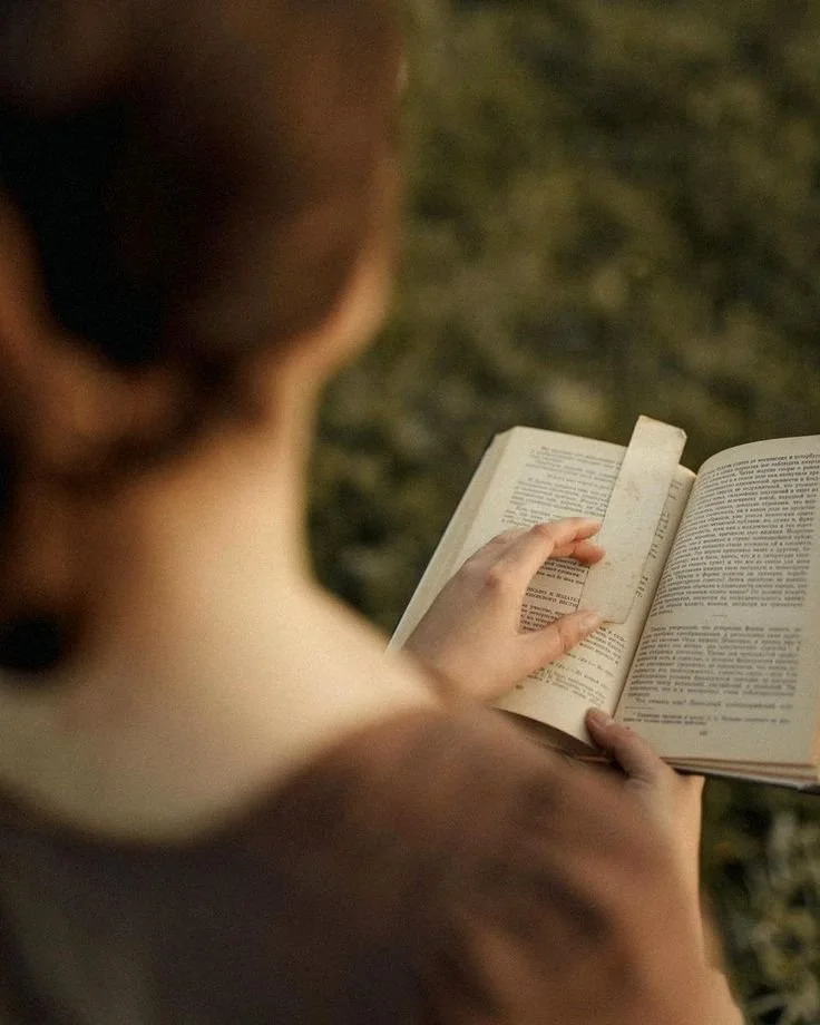 A person reading a book outdoors, with the photo taken from behind, showing the person's shoulder, neck, and hand holding the book.