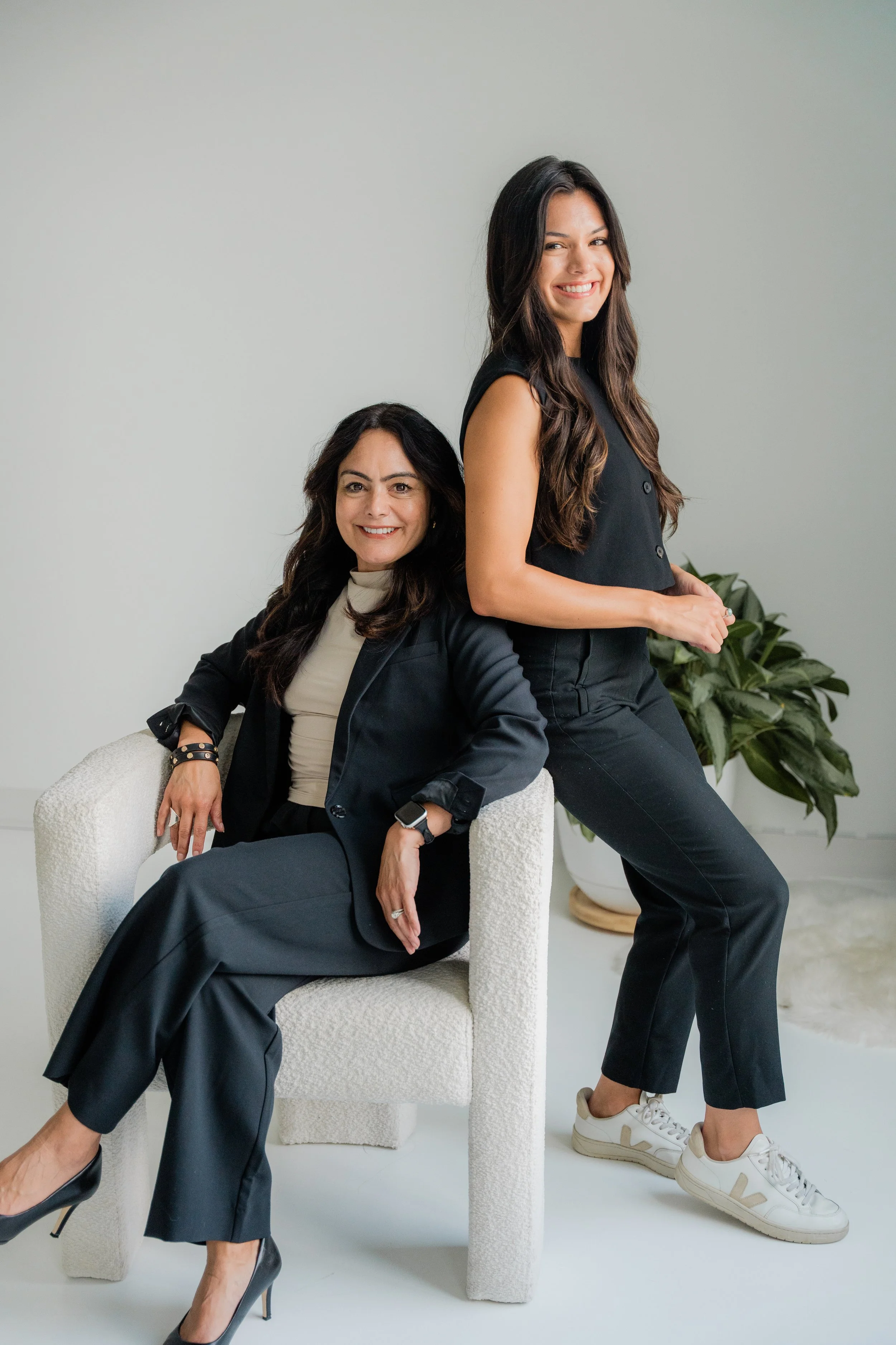 Two women dressed in black, one seated on a white textured armchair and the other standing beside her, smiling at the camera with a large plant behind them.