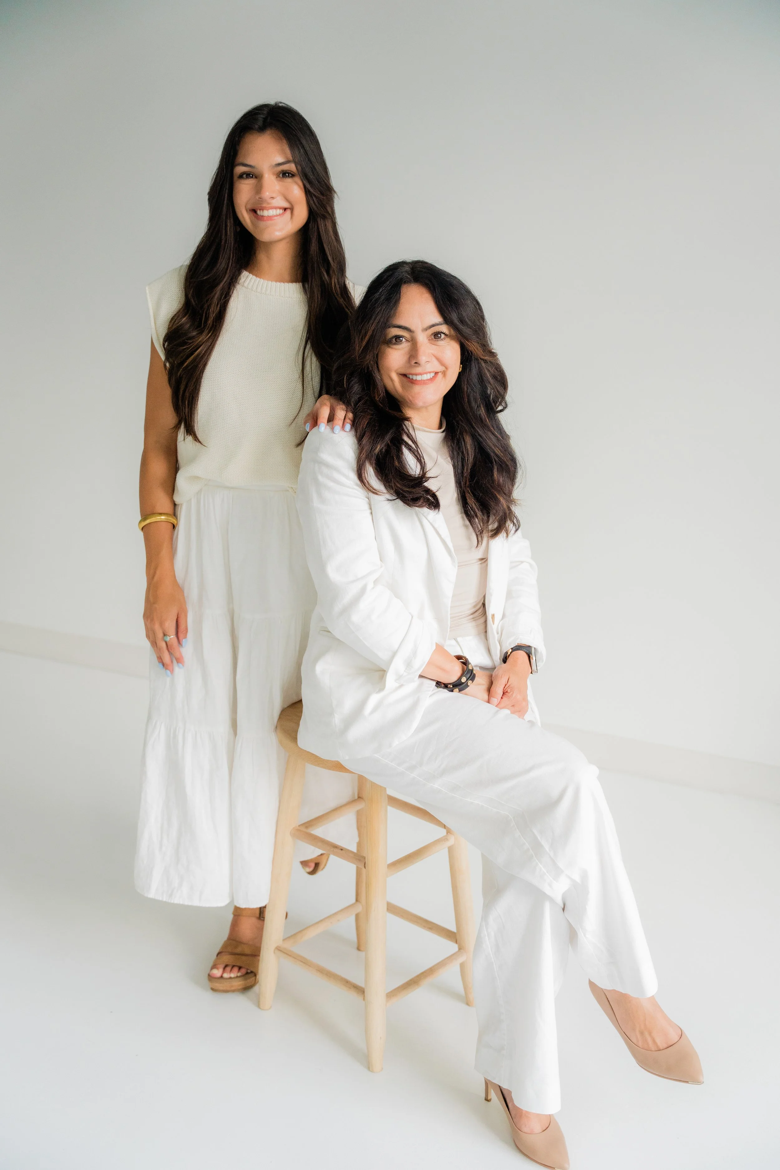 Two women, one sitting on a stool and the other standing behind her, smiling against a plain white background.