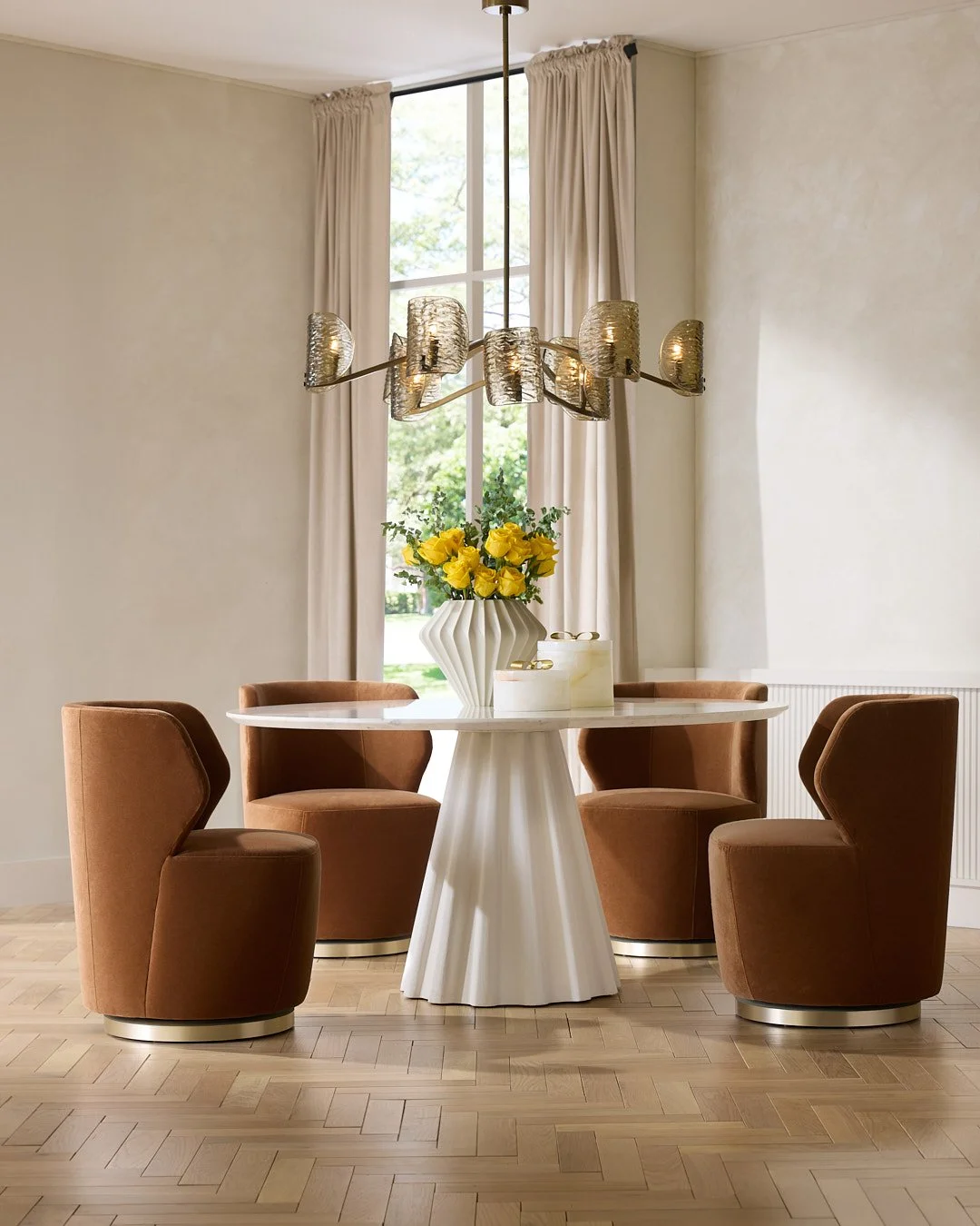 A round white dining table with a fluted column base, surrounded by four tan upholstered chairs. On the table is a white vase filled with yellow roses and green foliage. A modern chandelier with gold and textured glass shades hangs above the table. There is a large window with cream curtains behind, letting in natural light.