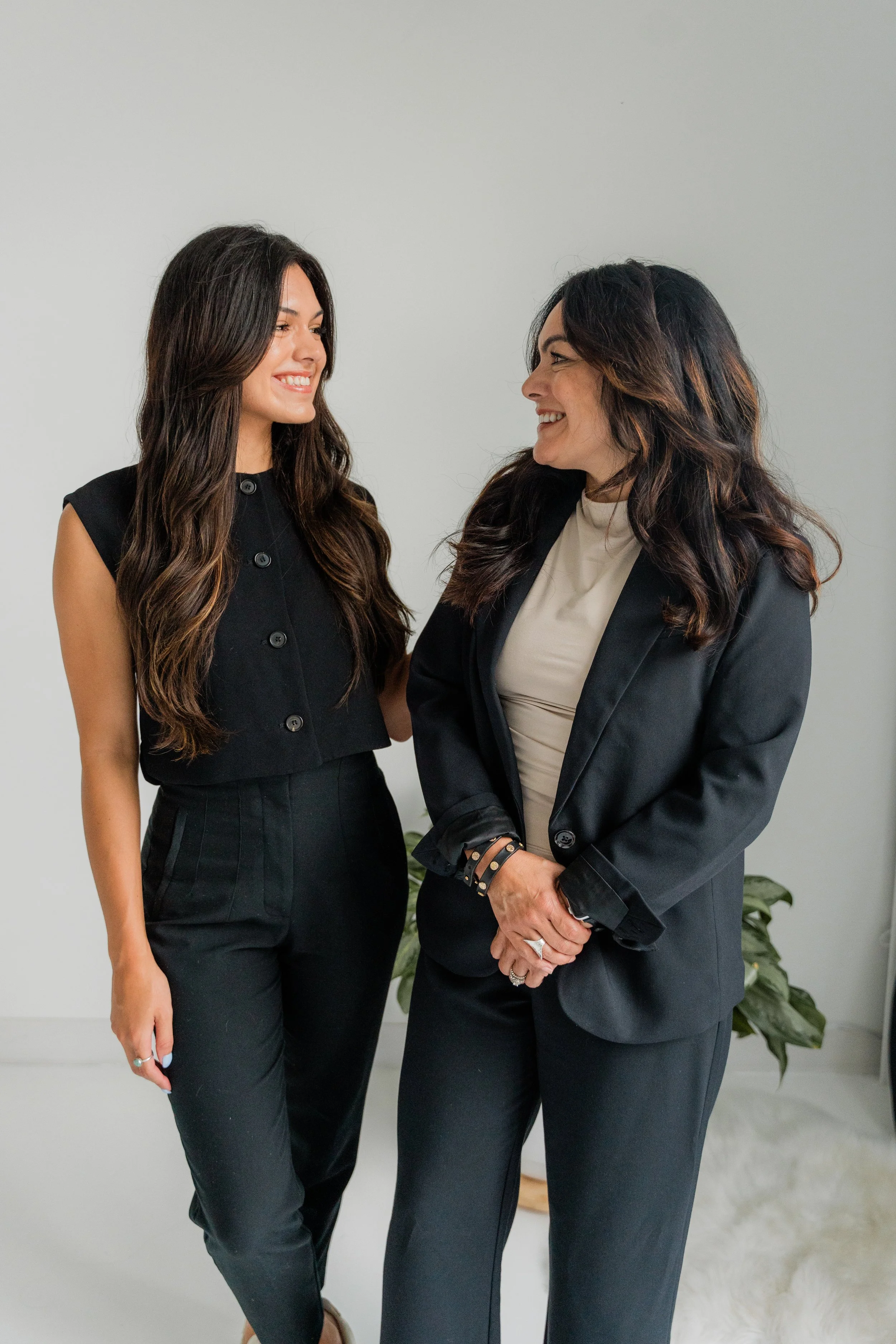 Two women smiling and looking at each other, dressed in black business attire, standing inside near a light-colored wall and potted plant.