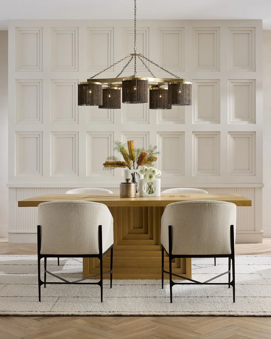 Modern dining room with a wooden table, four upholstered chairs with black metal legs, and a floral centerpiece in vases under a black and brass chandelier, against a white paneled wall.