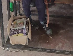 A person holding a hammer, standing near a box of snacks in a room with a concrete floor.