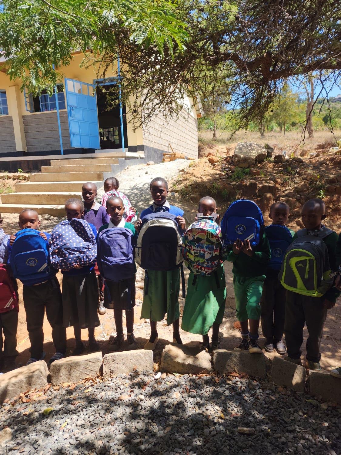 Group of school children standing outdoors in front of a building with stairs, holding backpacks, with trees and rocky terrain in the background.
